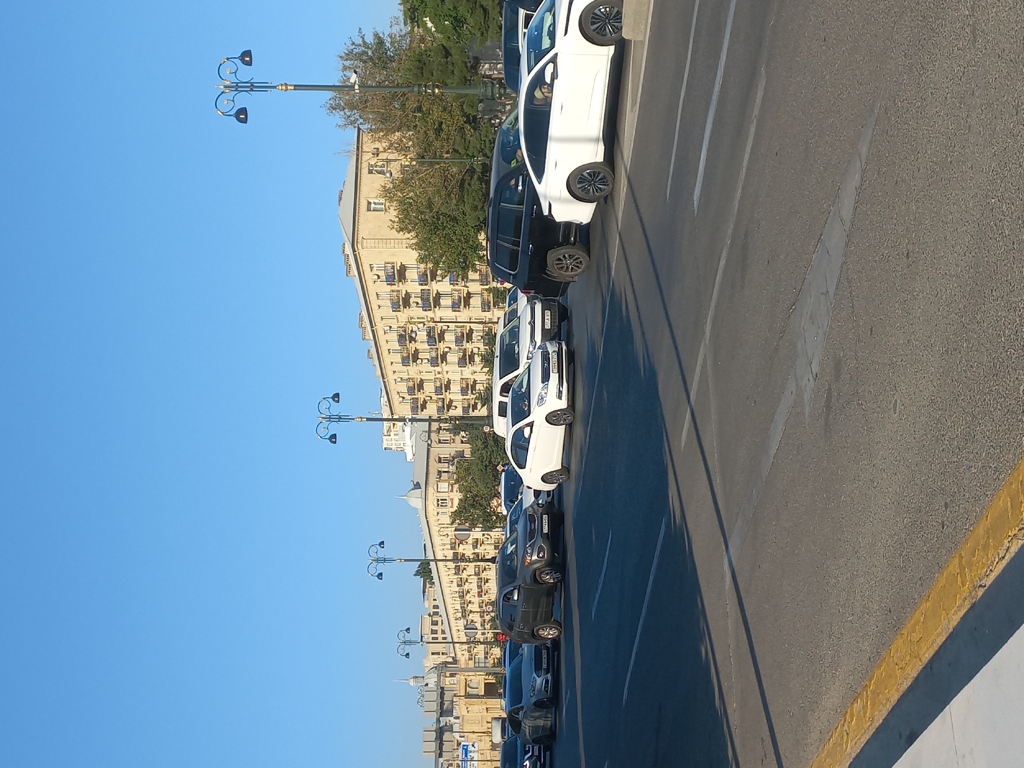 The image depicts a street scene in Baku, Azerbaijan, characterized by a clear blue sky and a row of buildings with ornate balconies and architectural details. Several vehicles are visible on the multi-lane road, indicating moderate traffic. The foreground shows the asphalt road with lane markings and a yellow curb. Streetlights with a distinctive design are positioned along the road. The buildings appear to be of a historic or classical style, with multiple stories and uniformed facades. Trees are present, adding greenery to the urban environment. The scene suggests a daytime setting with sunlight casting shadows. No people are clearly identifiable in the image, and the focus is on the urban landscape and vehicular traffic.