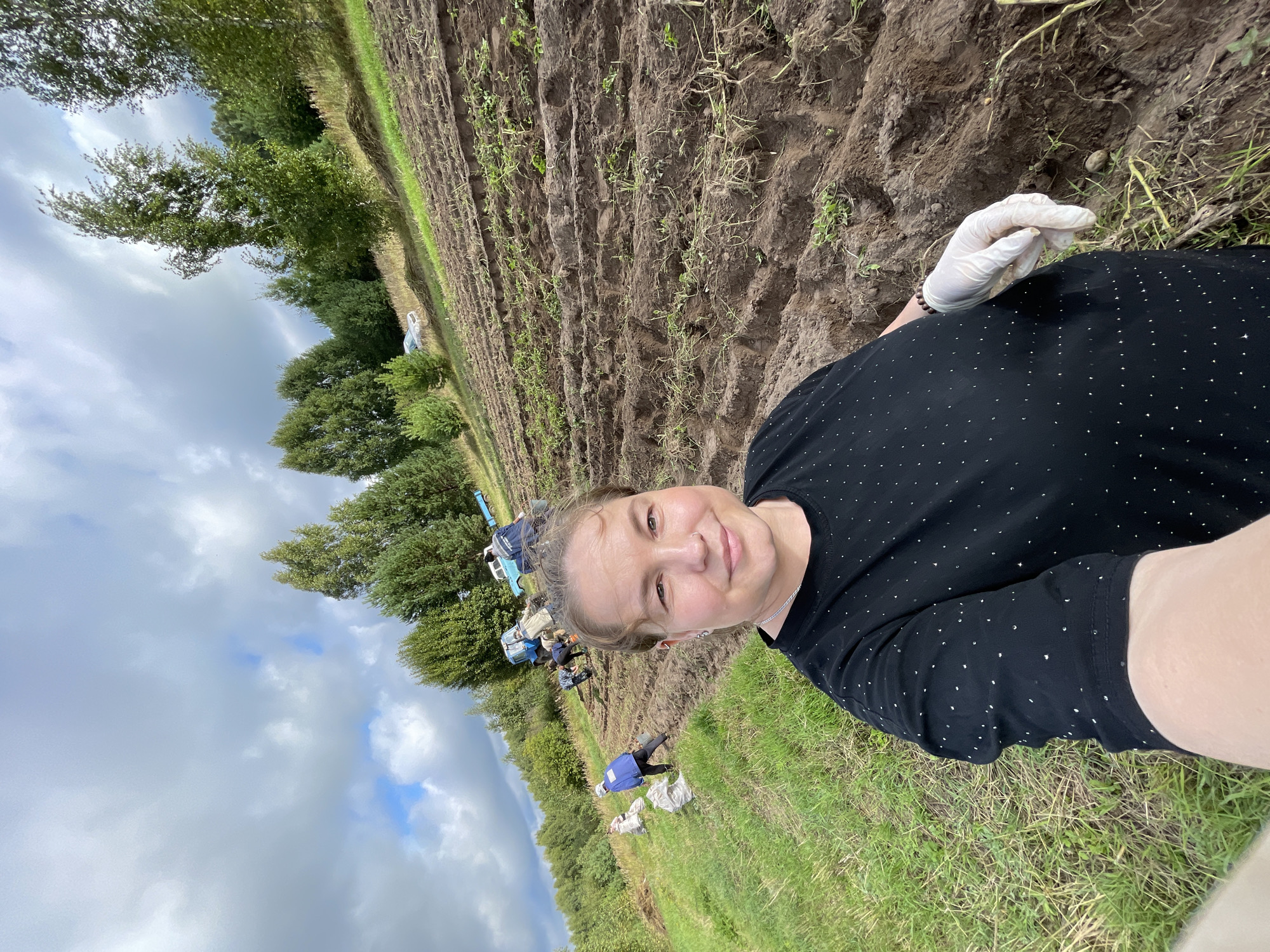 A woman in a black shirt with a silver necklace, wearing white gloves and a beaded bracelet, takes a selfie outdoors. She stands in the foreground, with a field of tilled soil and scattered green plants behind her. Several other people are working in the field further back, some near a blue tractor and a light-colored truck. Bags are visible on the left side of the field. The landscape is hilly, with trees and a cloudy sky. The location is identified as Baldelka, Russia.
