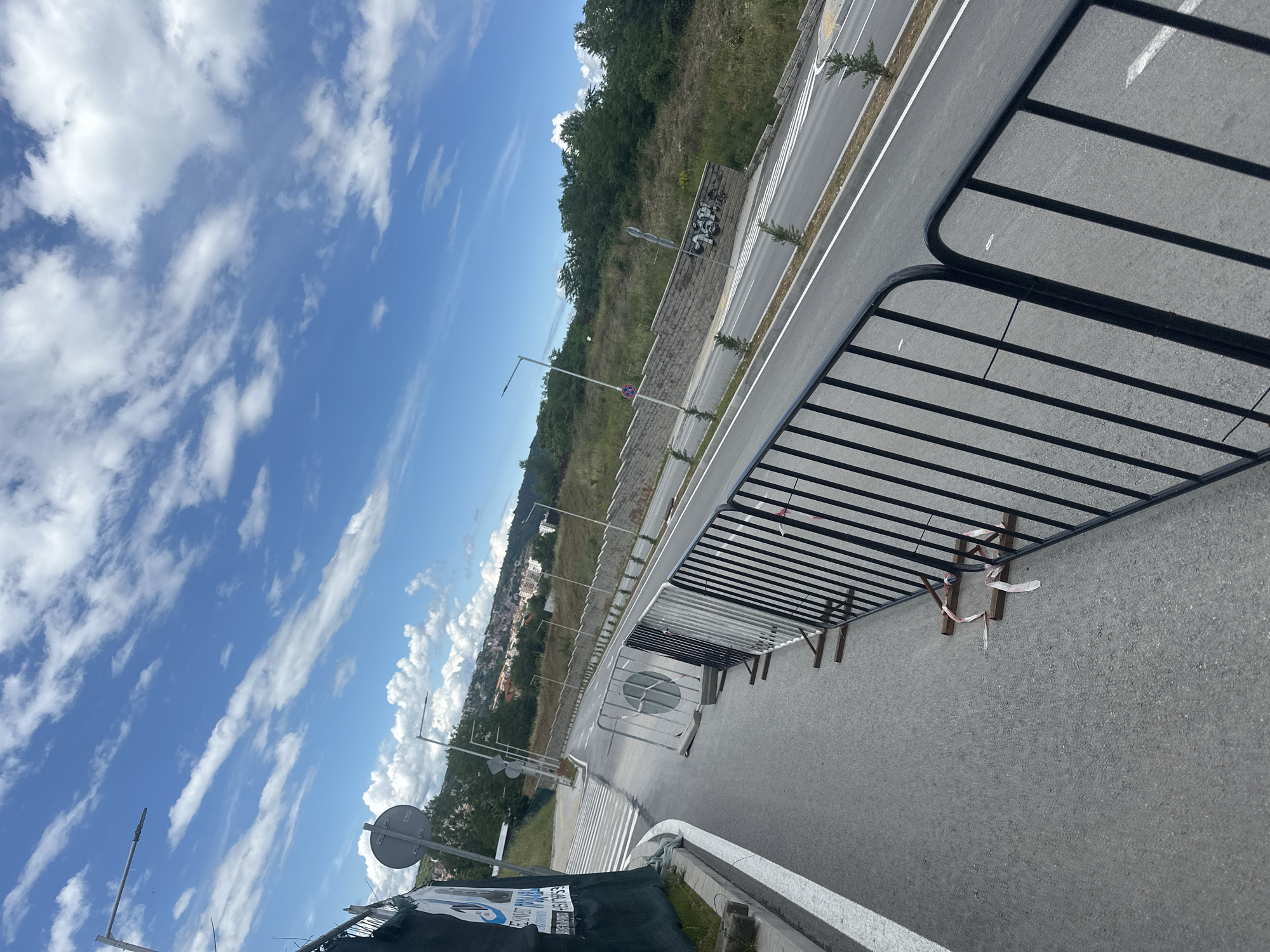 The image captures an outdoor scene of a modern road and its surroundings in Blagoevgrad, Bulgaria, under a bright, partly cloudy sky. The perspective is from an elevated viewpoint, looking along the road that stretches into the distance.
In the foreground and midground, a wide, recently paved asphalt road features clear white markings, including a pedestrian crossing and lane dividers. A prominent black metal temporary barrier system, typically used for crowd control or traffic management, runs along the right side of the road. Some segments of these barriers have red and white ribbons tied to them. On the left side of the road, additional barriers, some white, are visible, alongside streetlights and various road signs. One sign is a circular regulatory sign with a red border and a white bar, commonly indicating 'No Entry' or a similar restriction. Graffiti, including what appears to be numbers or letters like "1994", is visible on a stone retaining wall alongside the road.
Beyond the road, a verdant hillside covered with dense trees and grass rises. In the far distance, nestled among the hills, clusters of buildings can be seen, indicating an urban or suburban settlement.
The sky is a clear blue, adorned with numerous white, puffy cumulus clouds, suggesting fair and sunny weather. The bright illumination points to a daytime scene, likely morning or afternoon. There are no people or moving vehicles visible in the frame, suggesting the road might be newly constructed, under temporary closure, or awaiting activation.
At the bottom-left corner of the image, a partially visible banner or sign displays text fragments such as "ES MIKHAYLOV", "GRADNA", "OPOZHNITE", and "BLAGOVGRAD", the latter confirming the geographical context. Other fragmented words like "EVO" and "IZSHTAVANET" are also discernible.