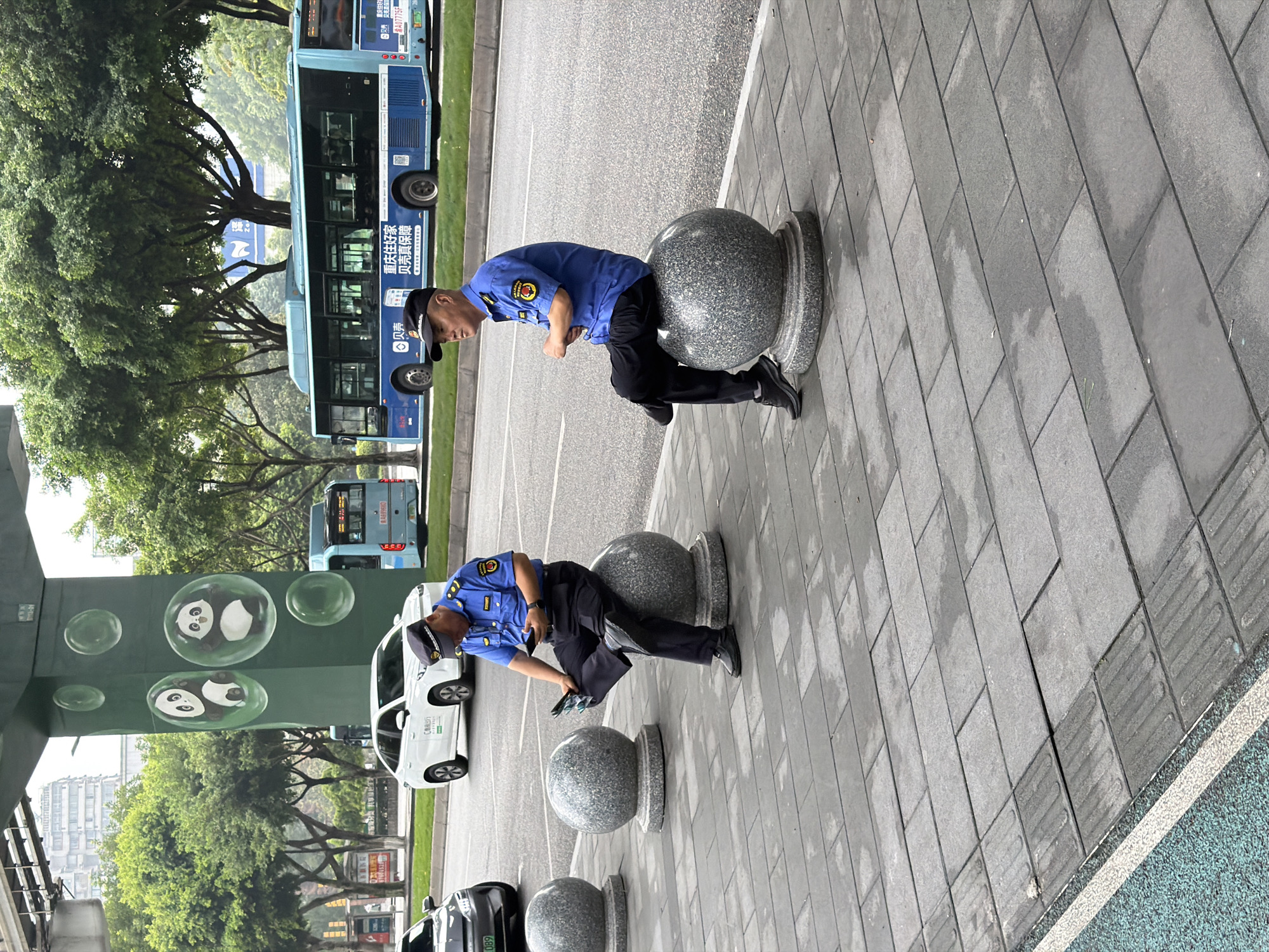 This image depicts a street scene in Jiulongpo District, China. Two men in blue uniforms and black caps are seated on decorative spherical stone pillars on a paved sidewalk. The man on the left is looking down at a mobile phone in his hand, while the man on the right is looking to his left, away from the camera. In the background, a blue city bus is visible on the road, along with a white car and other vehicles. A large green pillar adorned with painted panda figures and bubbles stands near the road. Trees line the street, and tall buildings are present in the distance. The ground is paved with grey rectangular tiles, and there are some wet patches, suggesting it may have recently rained.