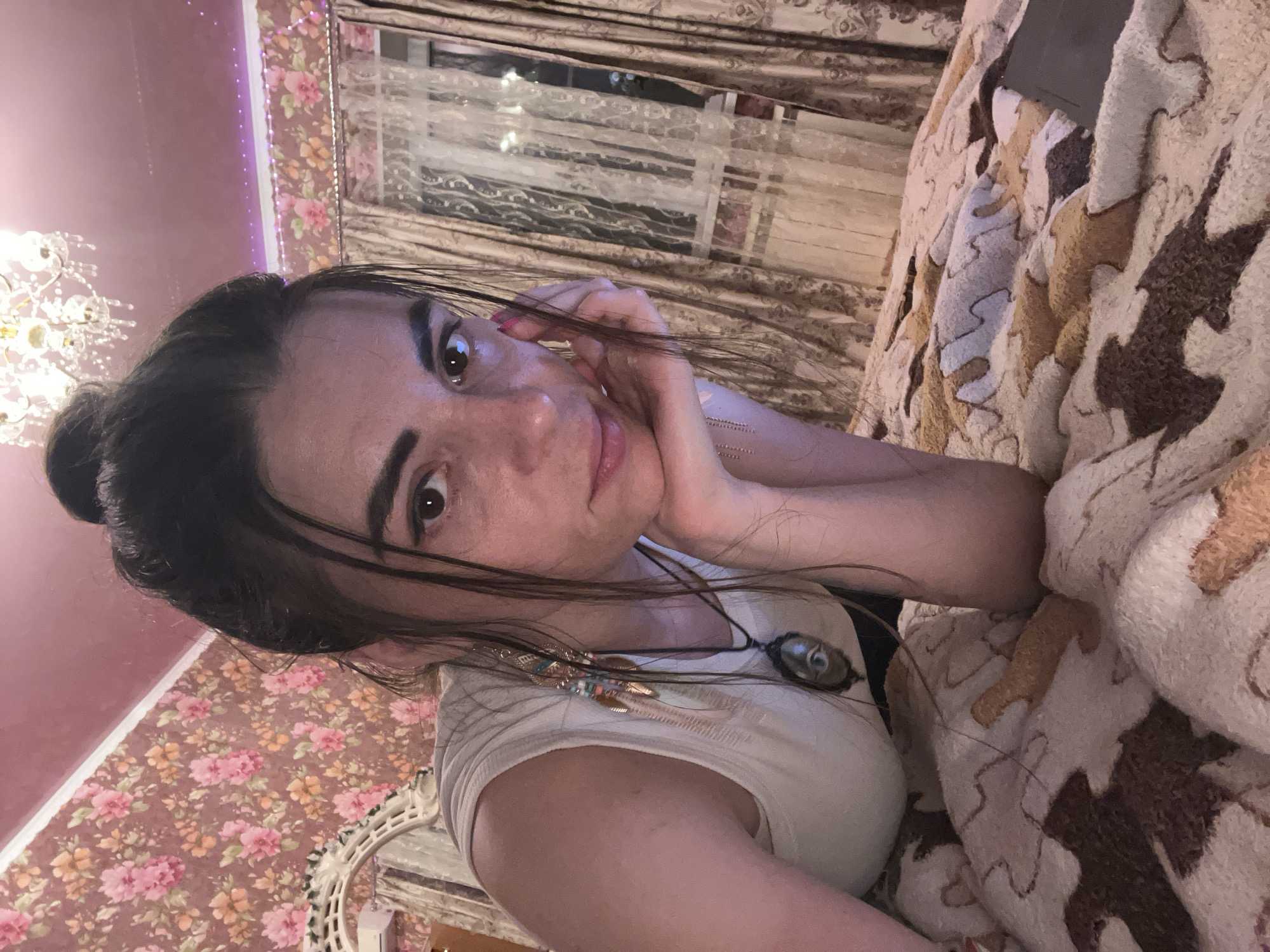 The image is a medium-shot photograph of a woman lying on a bed. She has dark hair tied up in a bun, and she is looking directly at the camera with her hand propped on her chin. She is wearing a white tank top and a pendant necklace with an eye-shaped charm. The bedspread has a patterned design with brown and white colors. In the background, the wall is decorated with floral wallpaper, and there are curtains over a window. A chandelier is visible on the ceiling. The overall setting appears to be a bedroom, likely in a residential location. The image does not provide any explicit information to pinpoint its location within the city of Krasnoe-na-Volge, Russia.