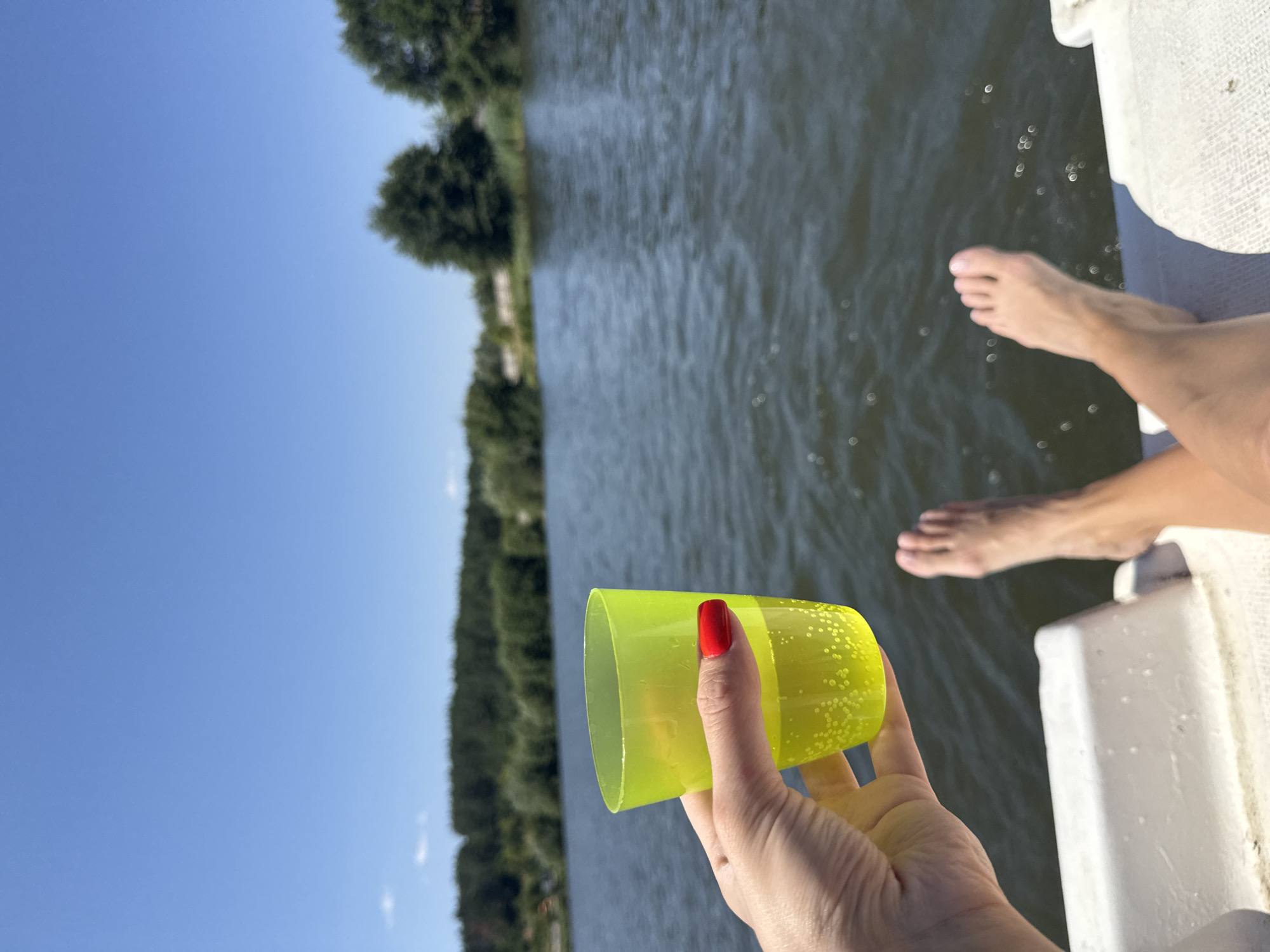 In Tiutky, Ukraine, on a bright, sunny day, a person is relaxing on a boat on a calm body of water. Their bare feet are visible, propped up on the edge of the boat. In their hand, they hold a neon yellow plastic cup filled with a bubbly, light-colored beverage. The background features a dense line of green trees bordering the water and a clear blue sky. The scene suggests a peaceful outdoor recreational activity.