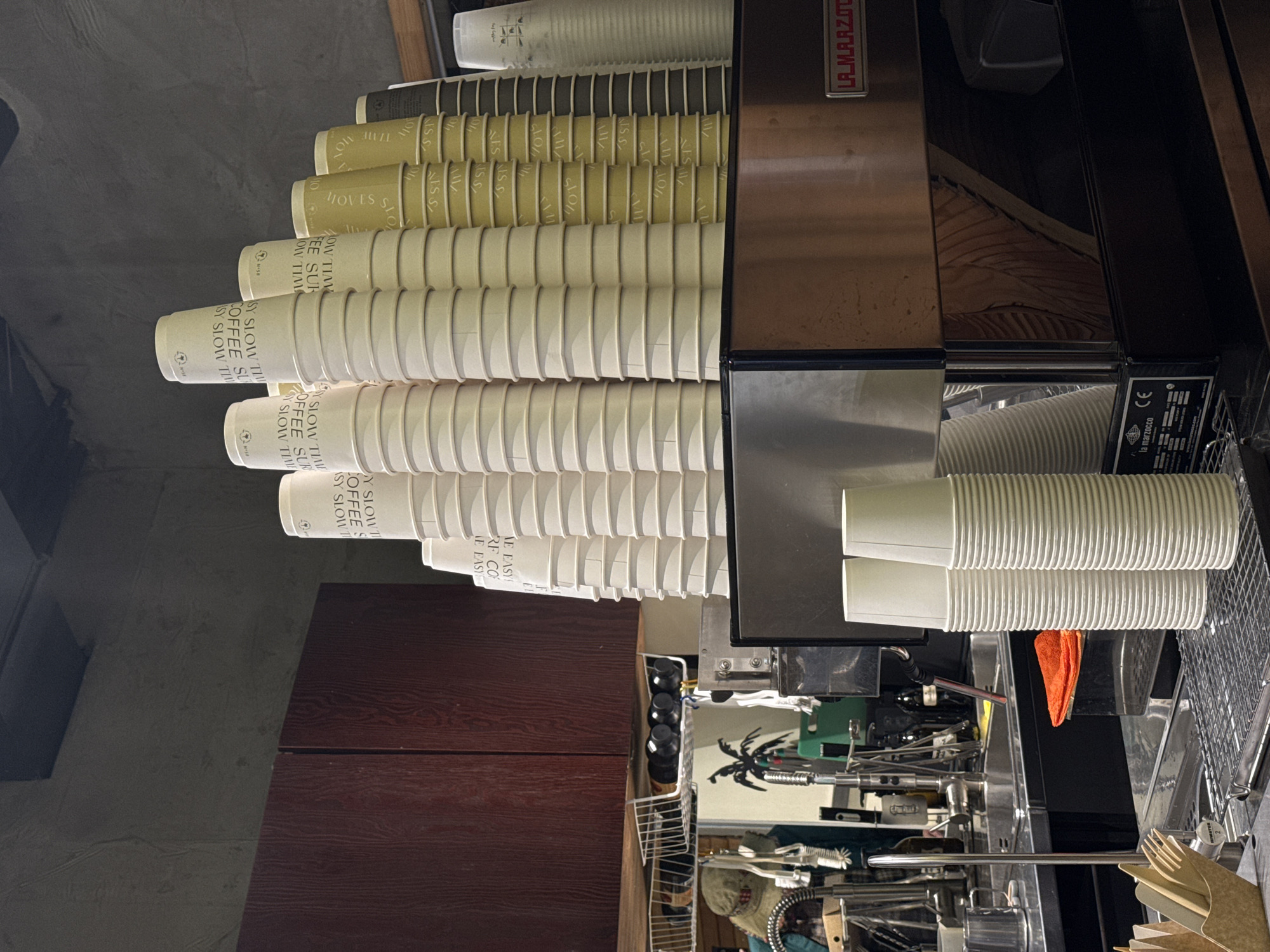 The image displays a close-up view of a coffee bar counter, likely within a cafe or shop. The scene is dominated by numerous, neatly stacked disposable coffee cups of various sizes and shades. Predominantly, stacks of cream-colored and light olive-tan cups are visible on the left and central parts of the frame.
On the right side of the counter stands a professional-grade coffee machine, bearing the branding "LA MARZOC" (likely La Marzocco) on its side and "la marzocco" along with a "CE" mark on a label below. Next to the machine, stacks of plain white paper cups are also present. The counter space around the coffee machine holds an assortment of coffee-making tools, a bright orange cloth, and a metal wire basket containing small dark bottles. Further to the right, a container of disposable wooden or bamboo cutlery can be seen.
No individuals are present in the image, suggesting a moment without active service. The setting conveys an organized and functional area for beverage preparation. Visible text on the coffee cups includes recurring phrases such as "SY SLOW TIME", "COFFEE SUP", "MOVES S", and variations like "COFFEE SY SLOW TIME". Some older or darker cups also show "NISS", "GLOI", and "110". This scene is located in Moscow, Russia.