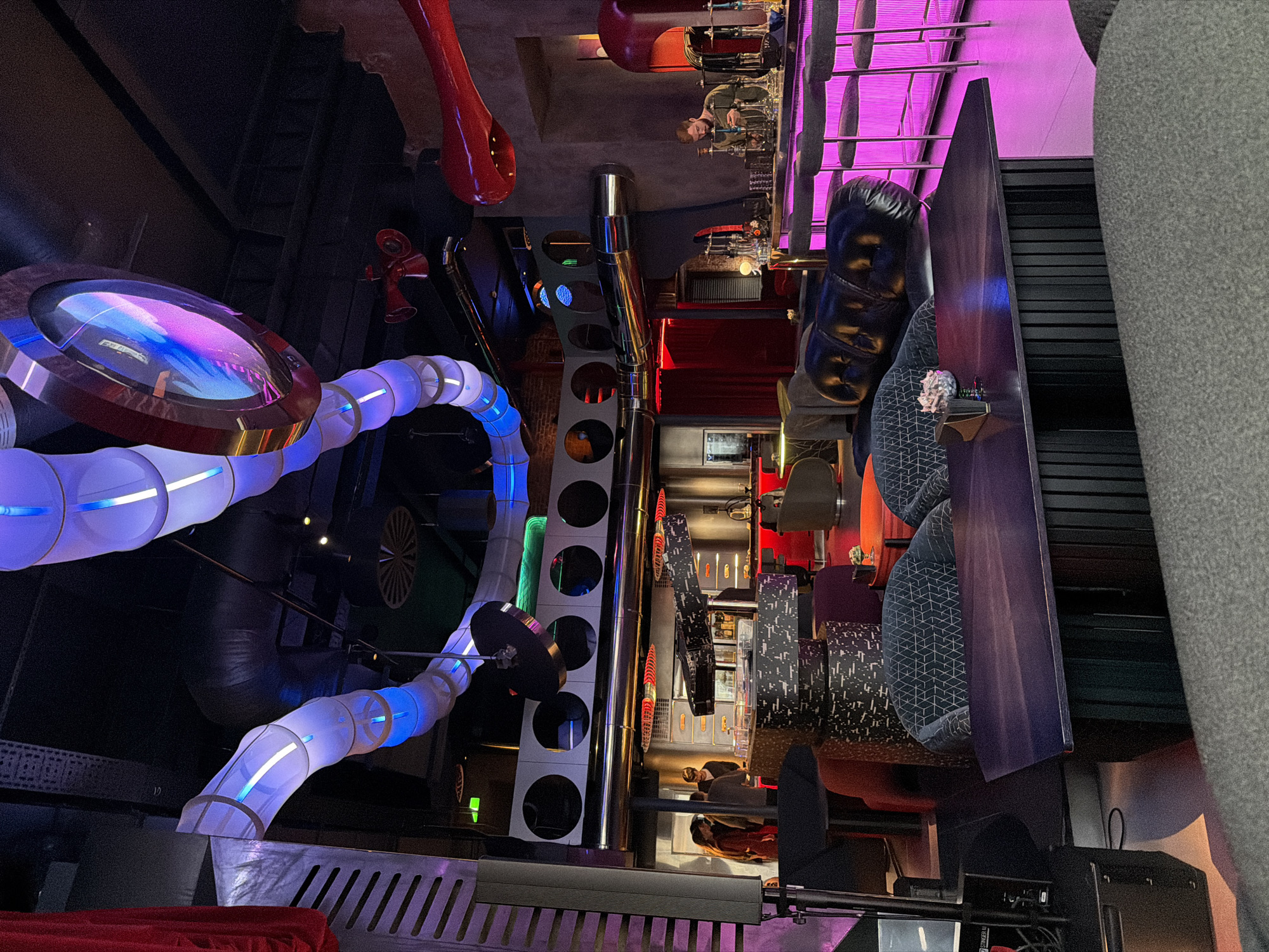 The image depicts the interior of a dimly lit, modern bar or restaurant with a distinct industrial and futuristic aesthetic, located in Moscow, Russia. The space is characterized by exposed metal pipes, ductwork, and circular cutouts in the walls, illuminated by vibrant blue, purple, and green LED lighting. Large, tubular light fixtures with internal blue lights hang from the ceiling, resembling futuristic conduits. A large, circular screen with a colorful, abstract display is suspended overhead.
In the foreground, dark wooden tables and plush, upholstered seating, including a black leather Chesterfield sofa, are arranged. Several patrons are visible in the background, including a bartender at a brightly lit pink bar, where customers are seated on stools. Another group of people are gathered near a service counter further in the background. The overall ambiance is chic and contemporary, suggesting a popular nightlife venue.