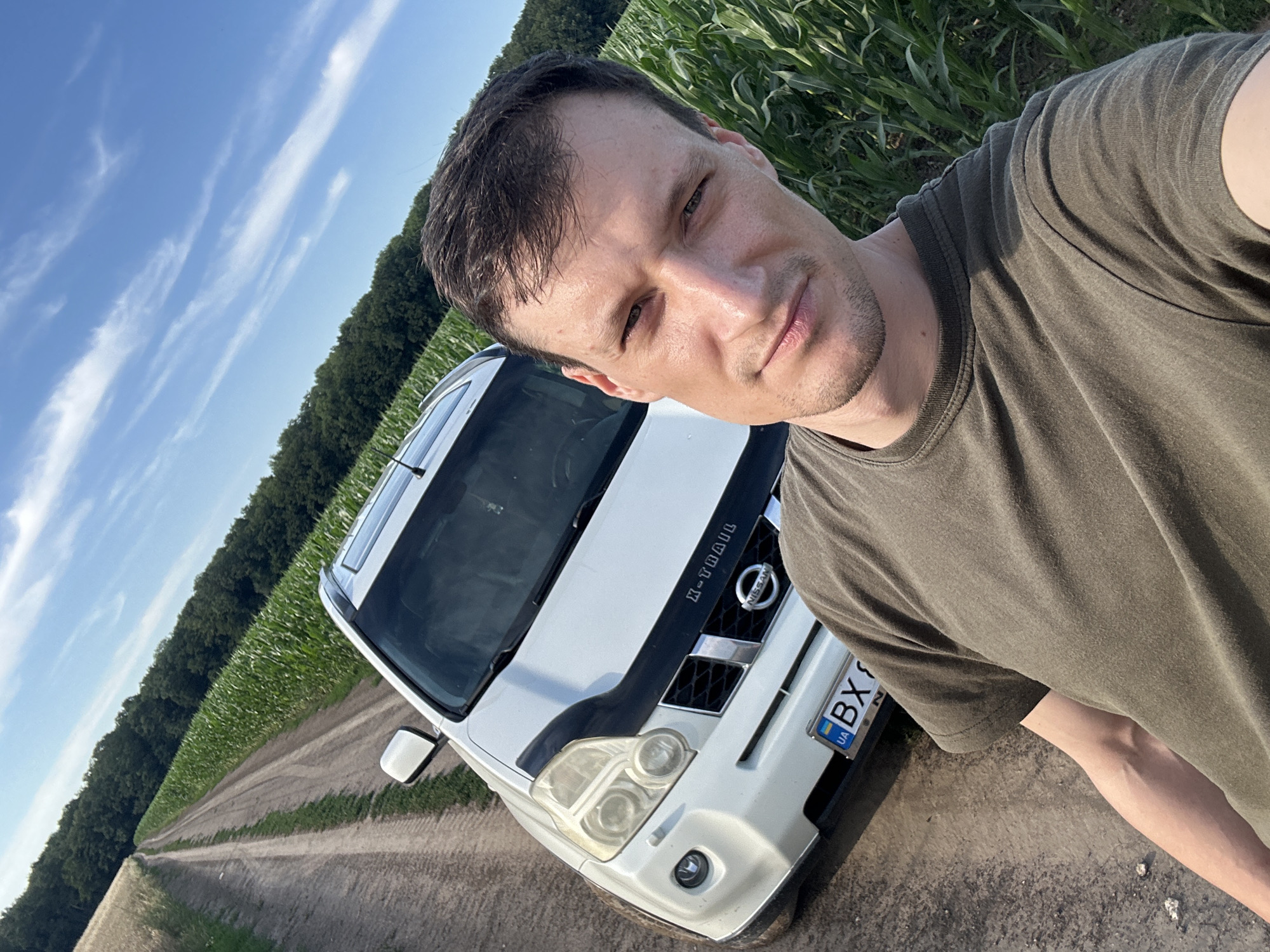 A man in a green t-shirt takes a selfie outdoors with a white Nissan X-Trail in the background. The car is parked on a dirt road beside a field of corn. The road extends into the distance, lined by trees on either side. The sky is blue with scattered white clouds. The license plate of the car reads "BX 9". The scene appears to be in or near Makiv, Ukraine, based on the license plate's format. The man has short brown hair and is looking directly at the camera.