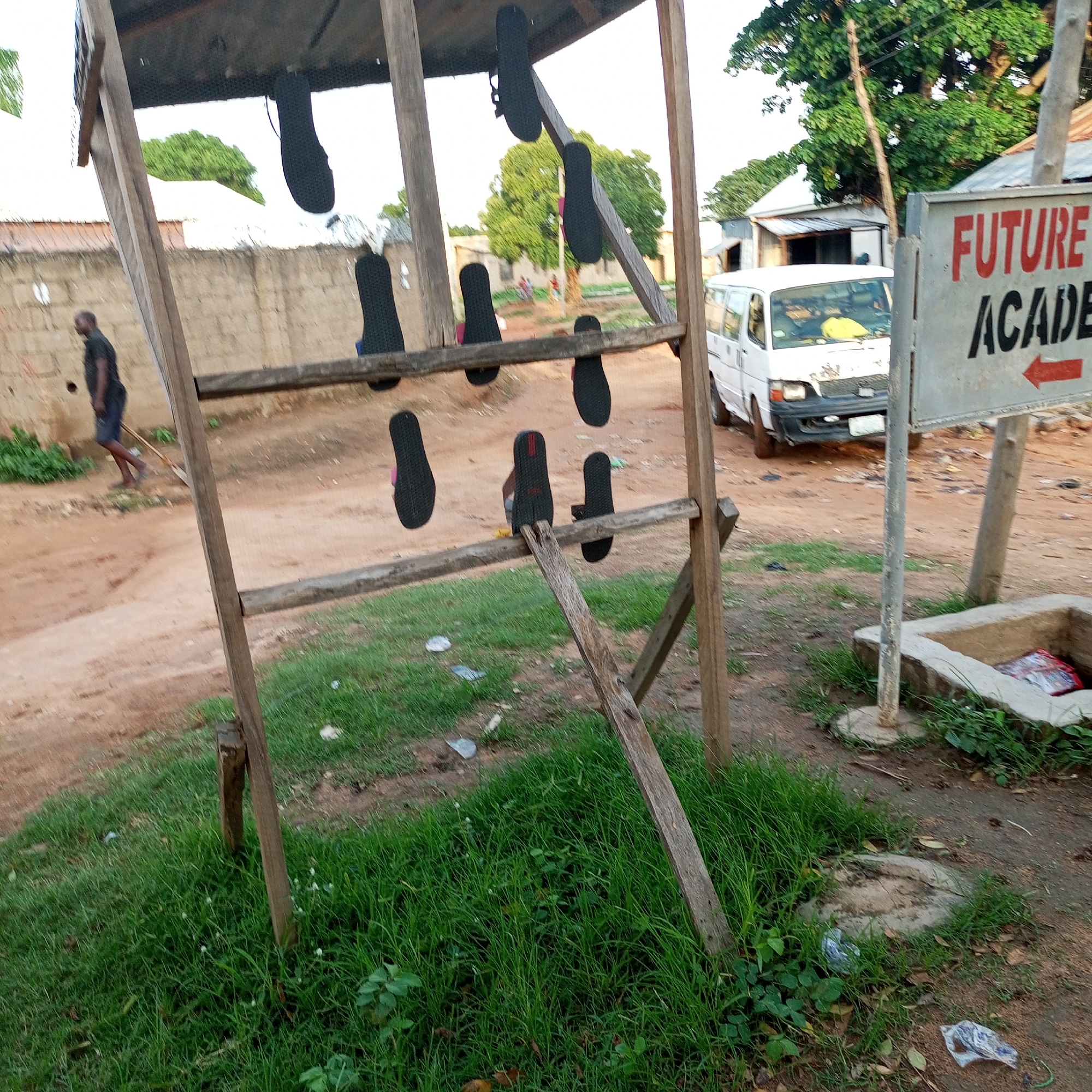 The image depicts an outdoor scene on a dirt road in Jalingo, Nigeria. In the foreground, a wooden stand displays several pairs of black flip-flops, some of which appear to be new and for sale. A white van is parked on the right side of the road, and a sign with the words "FUTURE ACADEMY" and an arrow pointing left is visible. A man is walking away from the camera on the left side of the road, and a beige brick wall lines the background. The environment appears to be a somewhat arid, dusty area with sparse vegetation, suggesting a rural or semi-urban setting.