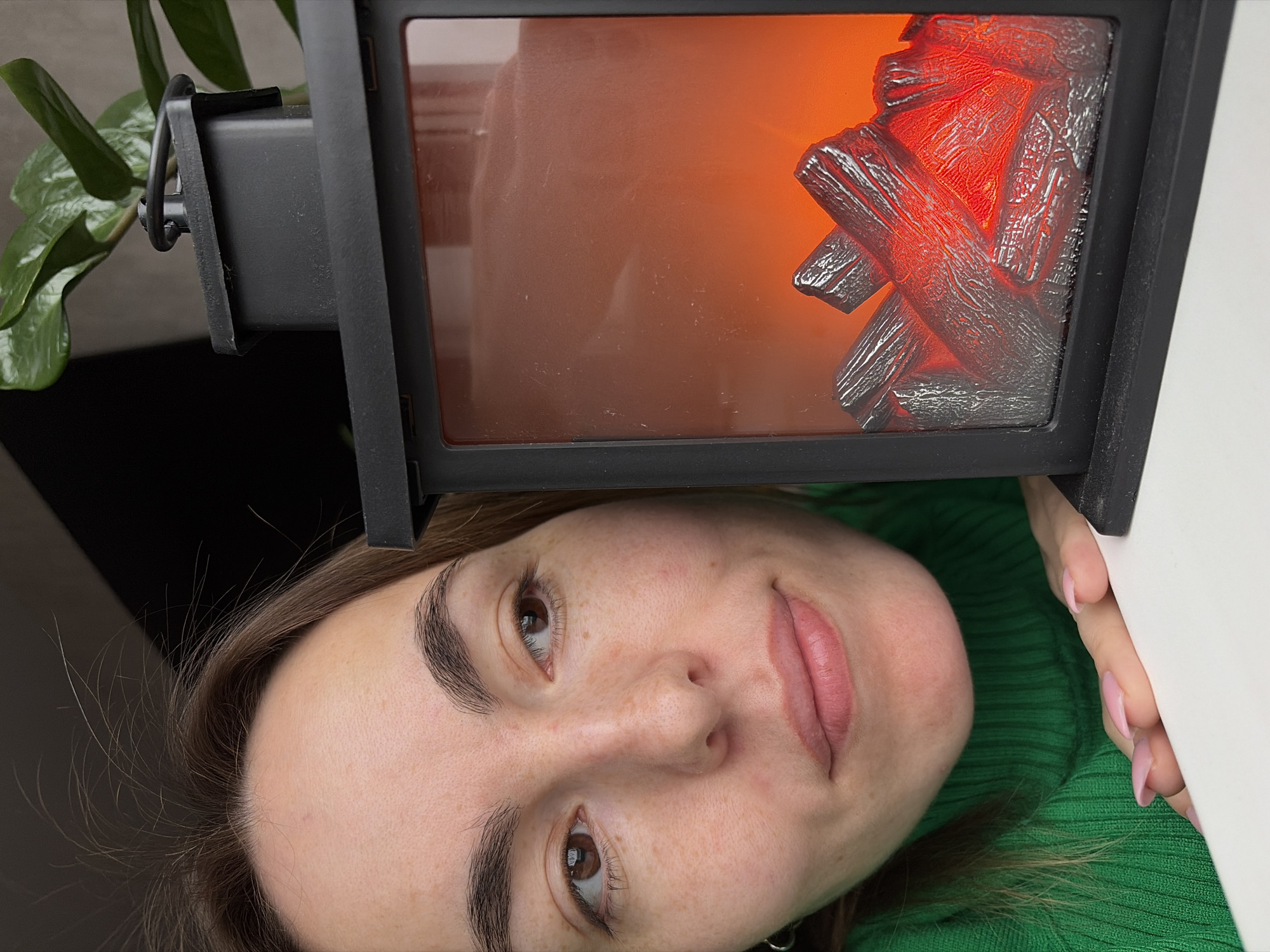 A close-up, upside-down perspective shows a woman's face positioned below a black frame that contains an artificial fire display. The woman has fair skin, brown hair, and brown eyes. She is wearing a green sweater. The black frame encloses a display that resembles burning logs, illuminated in orange and red tones. A green plant is visible on the left side, partially obscuring the black frame. A white surface is seen on the right. The location and city are not discernible from the image.