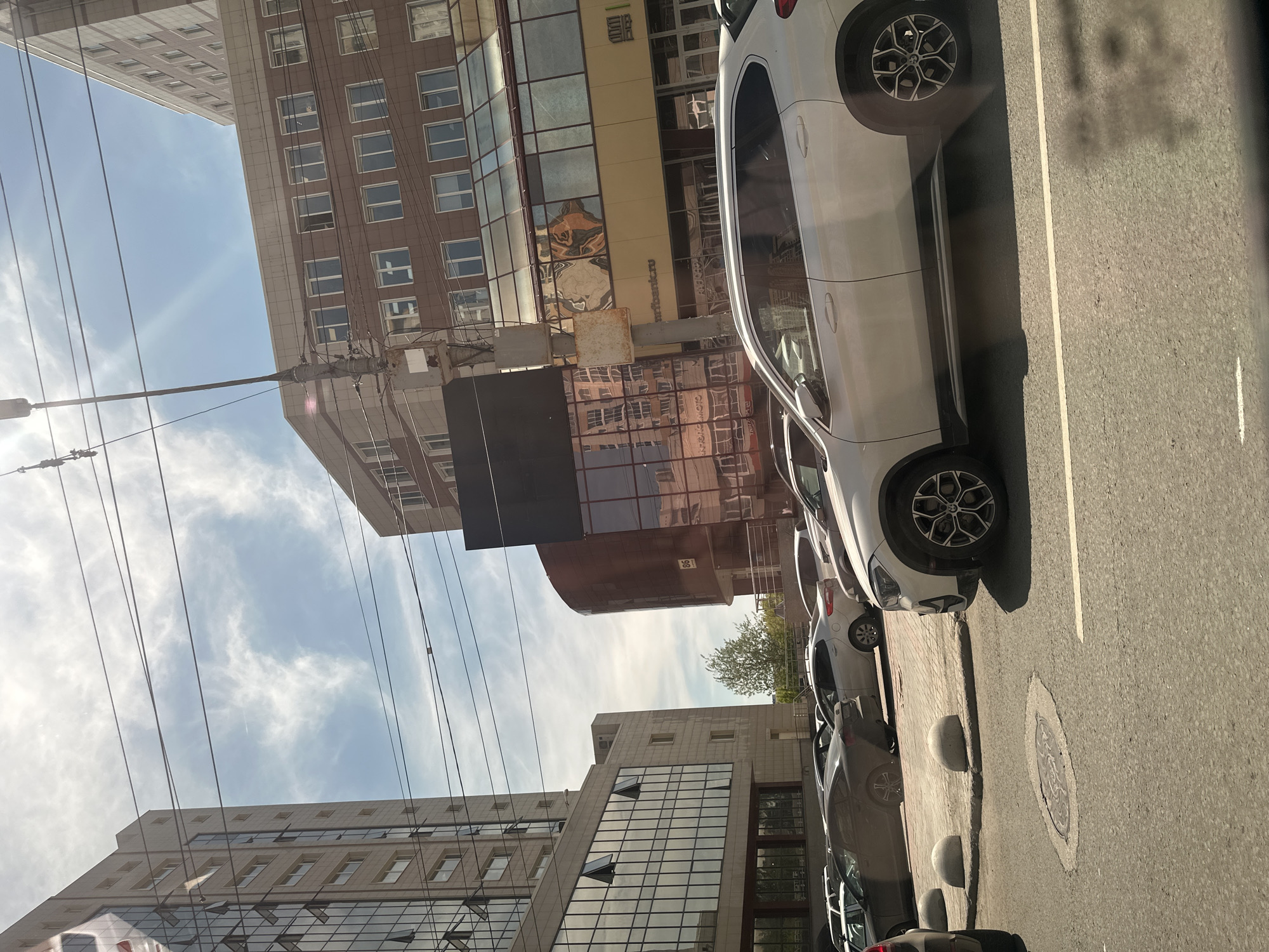 The image captures an urban street scene on a bright, partly cloudy day. A row of cars is parked along the right side of an asphalt road, which features a white dividing line. The closest vehicle is a white SUV or crossover with distinctive spoked wheels. The adjacent sidewalk has round concrete bollards and a manhole cover.
Multiple multi-story buildings dominate the background and left side of the frame. Some buildings feature modern glass facades, reflecting the sky and other structures, while others have more traditional designs with brown or beige paneling and numerous windows. On one building's facade, partial text reading "mrbankru" and what appears to be "БАНК" (Bank) in Cyrillic is visible. Another building displays the number "86".
The sky, visible above and between the buildings, is a bright blue with scattered white clouds, and a sun flare is present towards the upper left. Numerous dark overhead wires crisscross the sky. A small tree with green leaves is visible in the mid-ground, tucked between buildings. The scene depicts a typical city environment during daylight hours.
