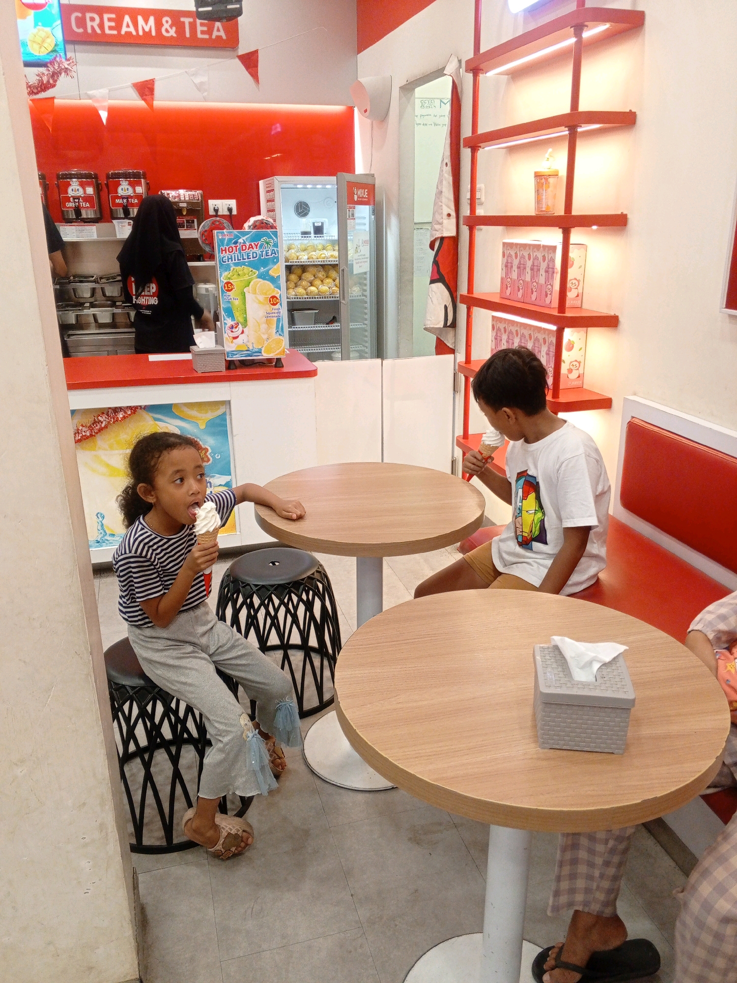 The image shows an indoor scene at a dessert shop in Gabus, Indonesia. Two children are present. A young girl with dark curly hair, wearing a striped t-shirt and grey pants, is seated on a black patterned stool and eating an ice cream cone. She is looking to her left. A young boy with short dark hair, wearing a white t-shirt with a graphic design and brown shorts, is seated on a red bench and eating an ice cream cone. He is looking to his left. In the background, a person wearing a black hijab and black t-shirt is working behind a counter. Red and white triangular flags are strung across the top of the counter area. A refrigerator displaying fruits and beverages is visible. Red shelving units with products are mounted on the wall to the right of the counter. Two wooden tables with white bases are in the foreground, one of which has a grey tissue box on it. The floor is tiled.