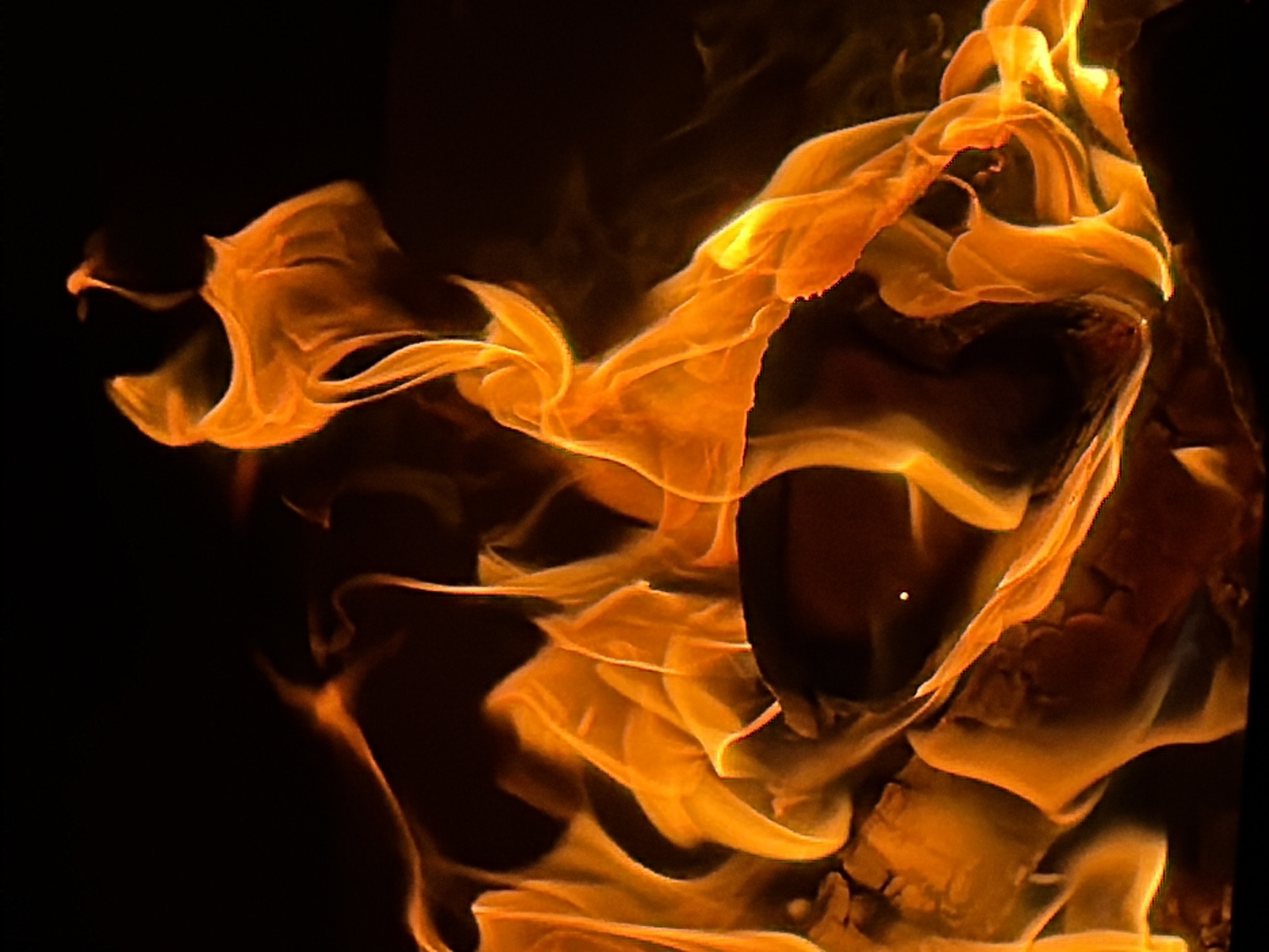 The image depicts a close-up view of intense orange flames against a black background. The flames are swirling and intricate, exhibiting varying shades of orange and yellow. There are no identifiable people, subjects, or objects other than the fire itself. The environment is dark, emphasizing the brightness of the flames. The location in the city of Zimyonki (Zimenki, Zimenka), Russia cannot be determined from the image.