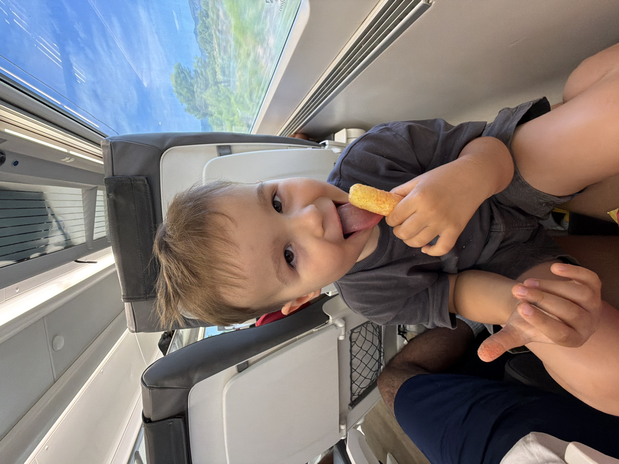 A young child, approximately two to three years old, is seated in a train carriage, looking directly at the camera with their tongue extended to lick a cheese puff. The child has light brown hair and is wearing a dark gray t-shirt and shorts. Their right hand holds the cheese puff, and their left hand is visible in the lower left quadrant of the image.
The train compartment appears to be a standard passenger carriage with gray upholstered seats. To the right of the child, a large window reveals a landscape of green hills and trees under a bright blue sky with scattered white clouds, suggesting daytime travel. The window shows streaks from the movement of the train, typical of a journey.
In the foreground, a portion of an adult's legs is visible, covered by dark blue shorts, indicating they are seated next to the child. The lighting suggests natural light coming from the window, supplemented by interior train lighting. The overall environment is that of a moving train, likely on a route that passes through scenic rural areas. There is no specific indication in the visual data that this scene is located in Tarragona, Spain, beyond the context provided in the prompt.