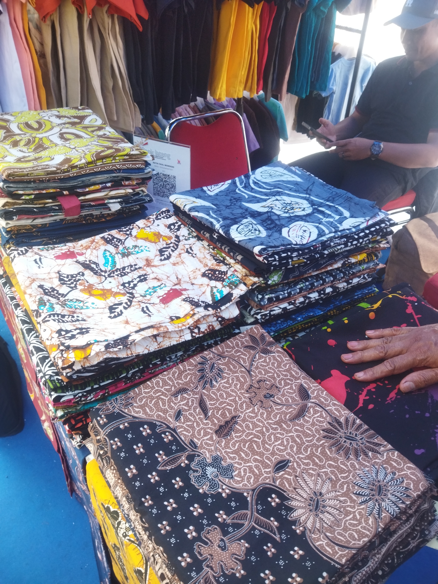 A stall displays stacks of batik fabric at a market in Giripurwo, Indonesia. The batik patterns are diverse, featuring floral motifs, abstract designs, and a butterfly-like print on a white background. A person, wearing a dark t-shirt and a baseball cap, is seated and holding a mobile phone. The background shows clothing racks with garments in various colors. The stall is set up outdoors, with a blue tarp covering the surface.