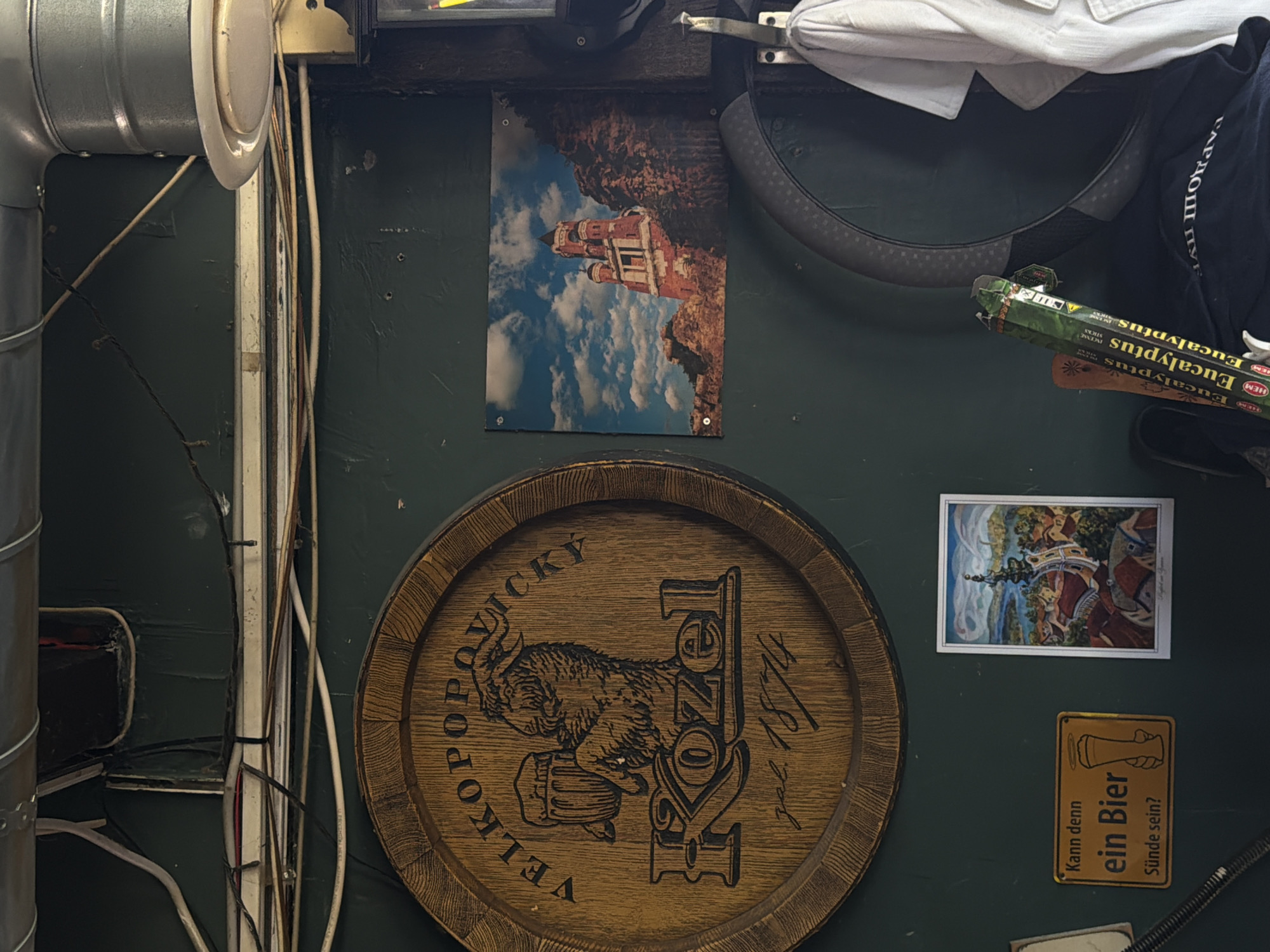 The image shows a cluttered interior scene with several items mounted on a dark green wall. On the left, a metal ventilation pipe runs vertically. Various electrical wires are haphazardly arranged. Central to the image is a wooden barrel-shaped sign with the inscription "VELKOPOPOVICKY KOZEL zal. 1874" and an image of an animal drinking from a mug. Above this, a small rectangular poster depicts a building atop a cliff against a blue sky. To the right, a partial view of a car steering wheel with a white garment draped over it is visible. Below the barrel sign, there is another small framed picture, and a yellow sign with the German text "Kann denn ein Bier Sünde sein?" A pack of HEM Eucalyptus incense sticks is partially visible. The general impression is of a somewhat disorganized storage or utility area. The city location of Palilula Urban Municipality, Serbia cannot be confirmed from this image alone.