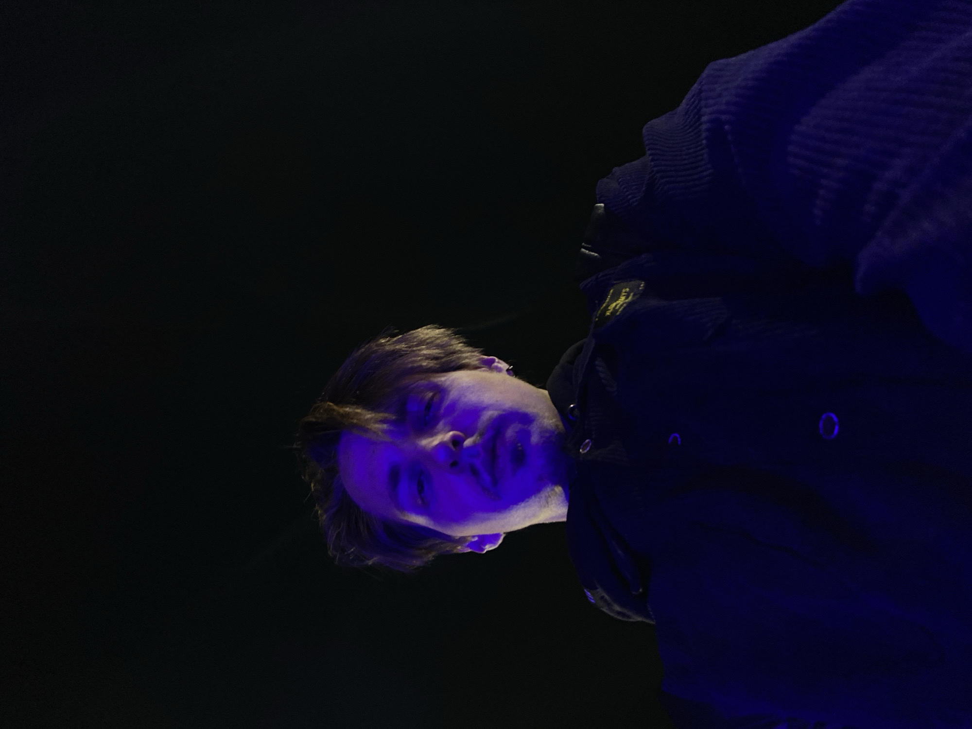 A low-angle shot, rotated counter-clockwise, captures a young man looking down towards the viewer against a solid black background. He is illuminated predominantly by a strong, cool purple or blue light, which casts a vibrant hue across his face, hands, and parts of his dark clothing, while leaving other areas in deep shadow.
The man has dark, medium-length hair and appears to have a neutral or slightly contemplative expression. He is wearing a dark, possibly black, button-up jacket or coat. On his upper left arm, there is a rectangular patch that appears to be yellow or gold with dark, illegible text or a logo. Beneath this jacket, the ribbed texture of another dark blue garment, possibly a sweater or hoodie, is visible, particularly around his right shoulder and arm.
The deep shadows and vibrant artificial lighting suggest an indoor setting, likely a venue such as a bar, club, or concert hall, consistent with the given location context of Bar, Montenegro. The time of day is presumably night. The photo captures a moment of observation or interaction, with the man's direct gaze drawing the viewer in. No other subjects or objects are clearly visible, emphasizing the man and the dramatic lighting.