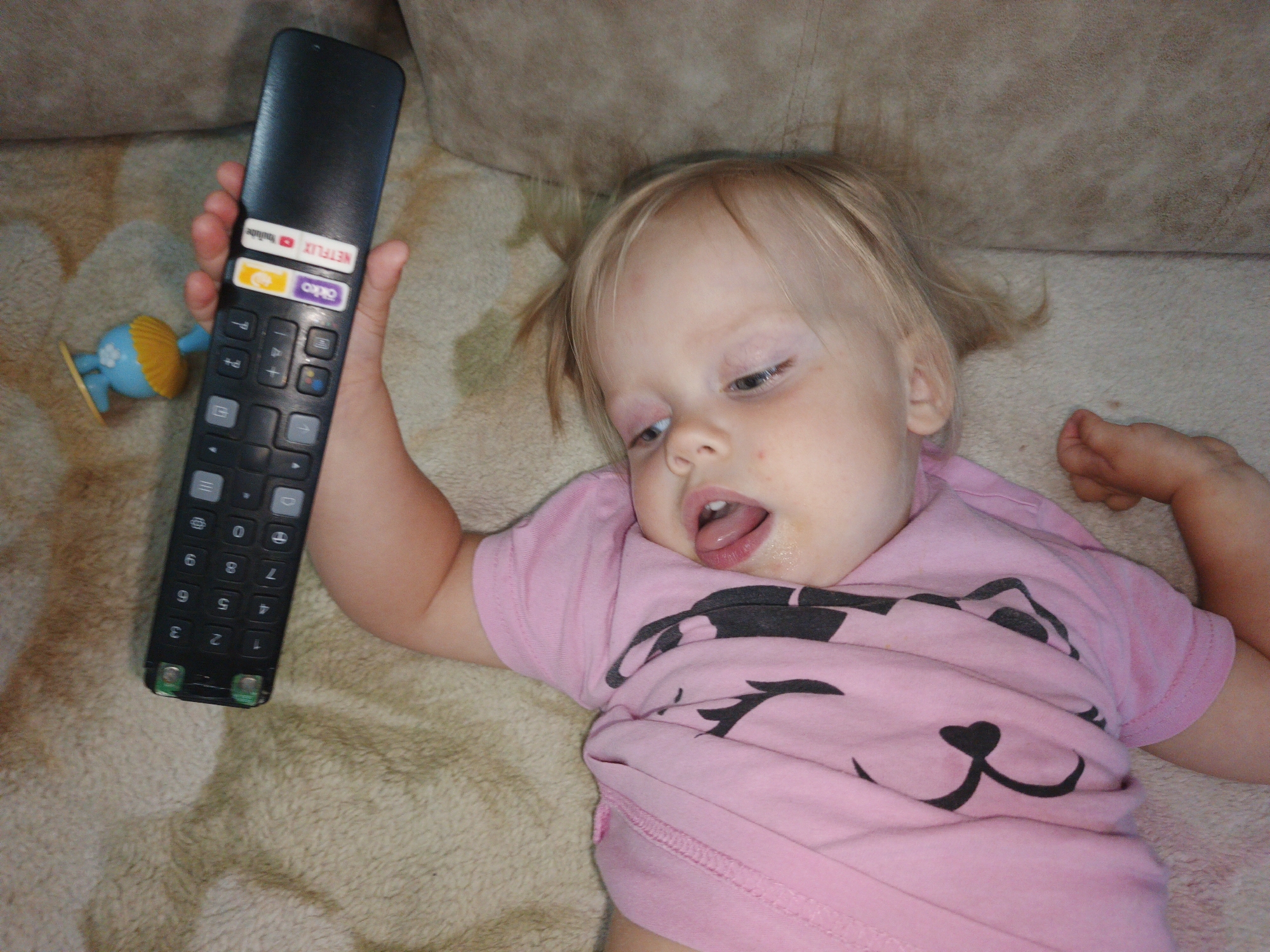 A close-up shot captures a toddler lying on a textured surface, possibly a blanket or carpet, with a couch in the background. The toddler, wearing a pink t-shirt with a black animal face graphic, has blonde hair and is looking towards the right side of the frame with their mouth open, revealing their tongue. A black remote control, held in the toddler's right hand, is positioned vertically in the image, displaying buttons and labels such as "NETFLIX" and "YouTube." To the left of the remote, a small blue and yellow toy is visible. The overall scene is brightly lit, with no specific identifiable location in Zlatoust, Russia.