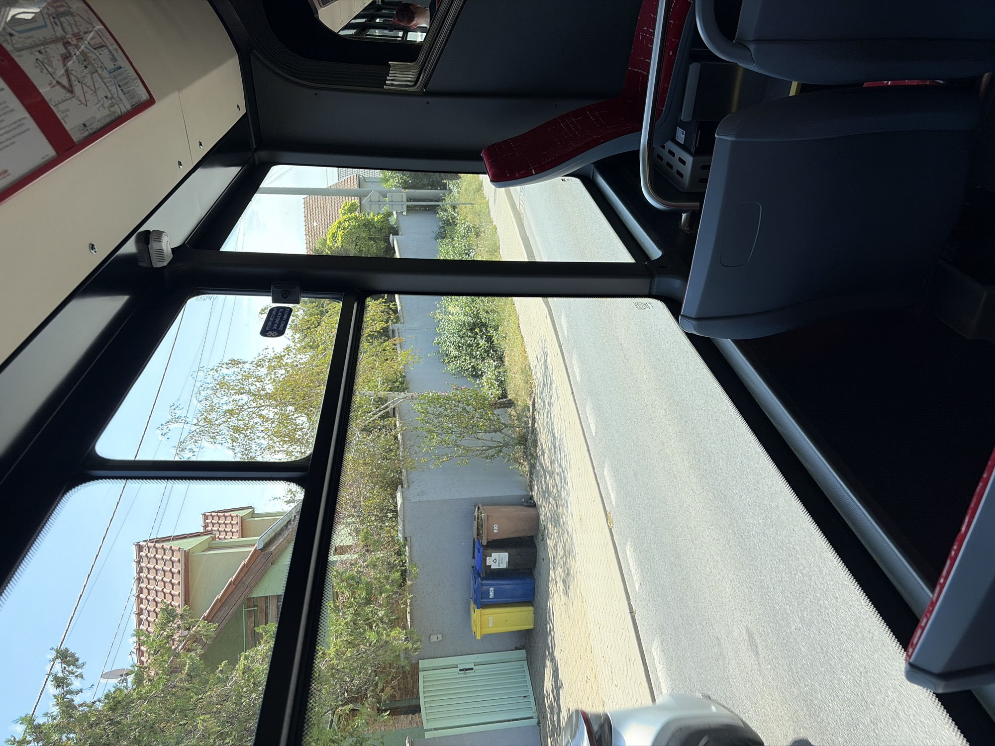 The image is taken from inside a bus, looking out the window at a street scene in Nitra, Slovakia. The window frames a view of a residential area with houses, a fence, and a row of four colored garbage bins on a sidewalk. The bins are yellow, blue, black, and brown. The street itself is paved and appears to be a residential road. The bus interior is visible in the foreground, with dark-colored seats and red accents on one of the seats. The sky is blue and clear.