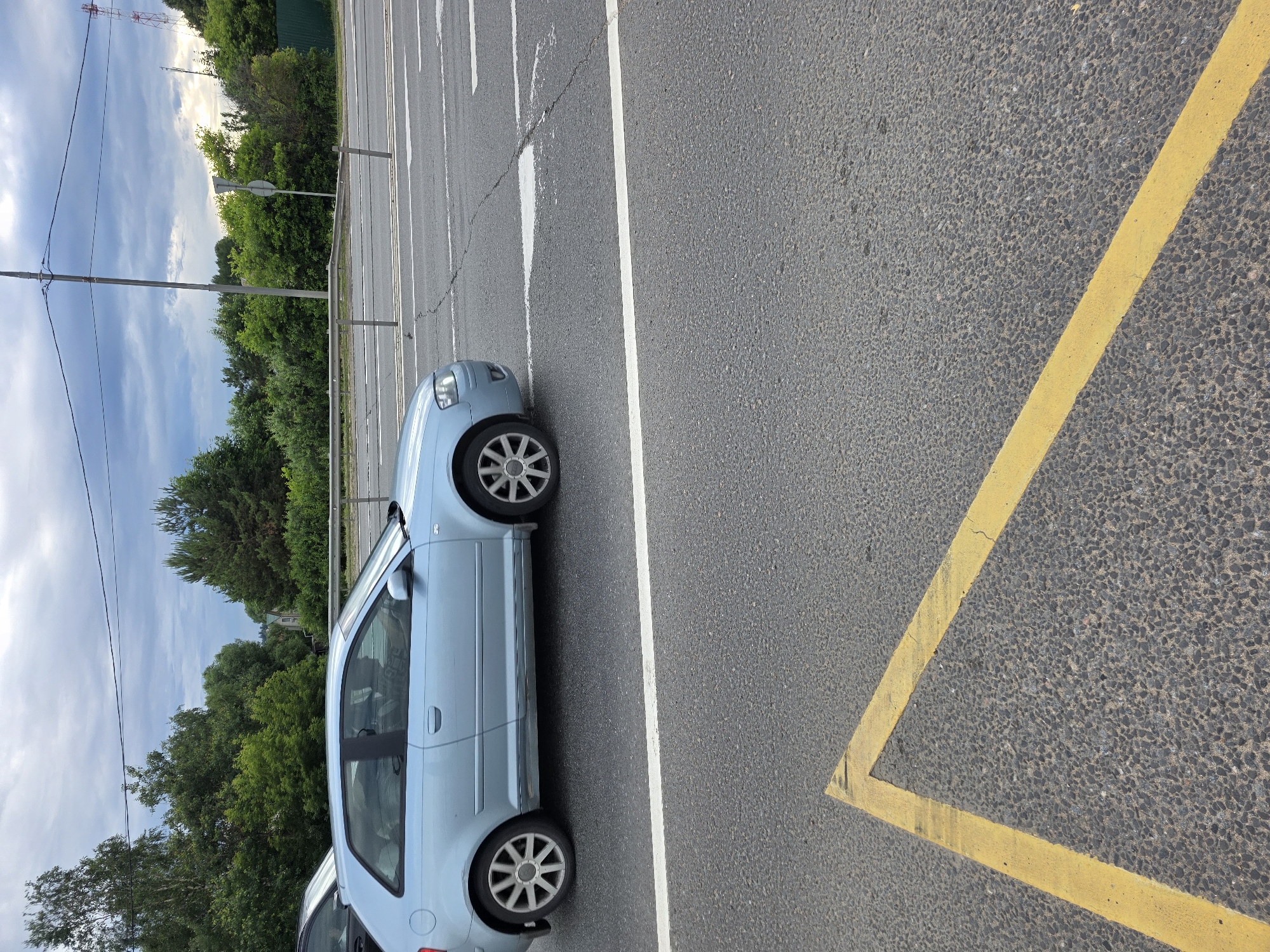 An outdoor, medium-shot, eye-level, vertical photograph captures a light blue, three-door hatchback car driving on a paved road. The car is angled to the left, with its front tires visible. The car's windows are tinted, obscuring a clear view of the interior. A person, likely the driver, is partially visible through the driver's side window. The road is marked with a white line on the left and a yellow line forming a triangle on the right. The background features a lush green forest, a grey metal guardrail, a utility pole, a circular traffic sign, and a tower on the right side. The sky is overcast with dramatic clouds. The ground is paved asphalt with visible cracks.