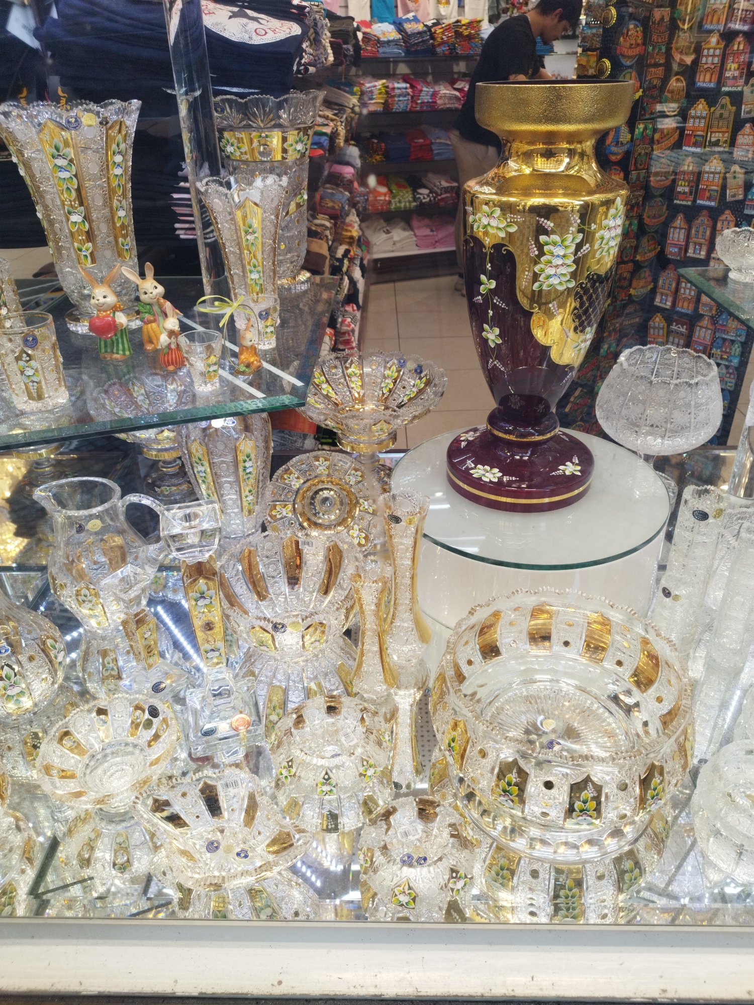 This is a photograph of a display of decorative glassware, likely taken inside a shop in Prague, Czechia. The items are arranged on mirrored surfaces, creating reflections. Prominently featured are several ornate crystal vases and bowls, many with gold accents and painted floral designs in shades of green, white, and blue. Among the glassware are small ceramic figurines, including two rabbits holding an apple and another holding a small bird. In the background, shelves are visible stocked with t-shirts and other merchandise. A man wearing a black shirt is partially visible in the background, appearing to be a shop employee or customer. The overall impression is one of a retail environment selling decorative and souvenir items.
