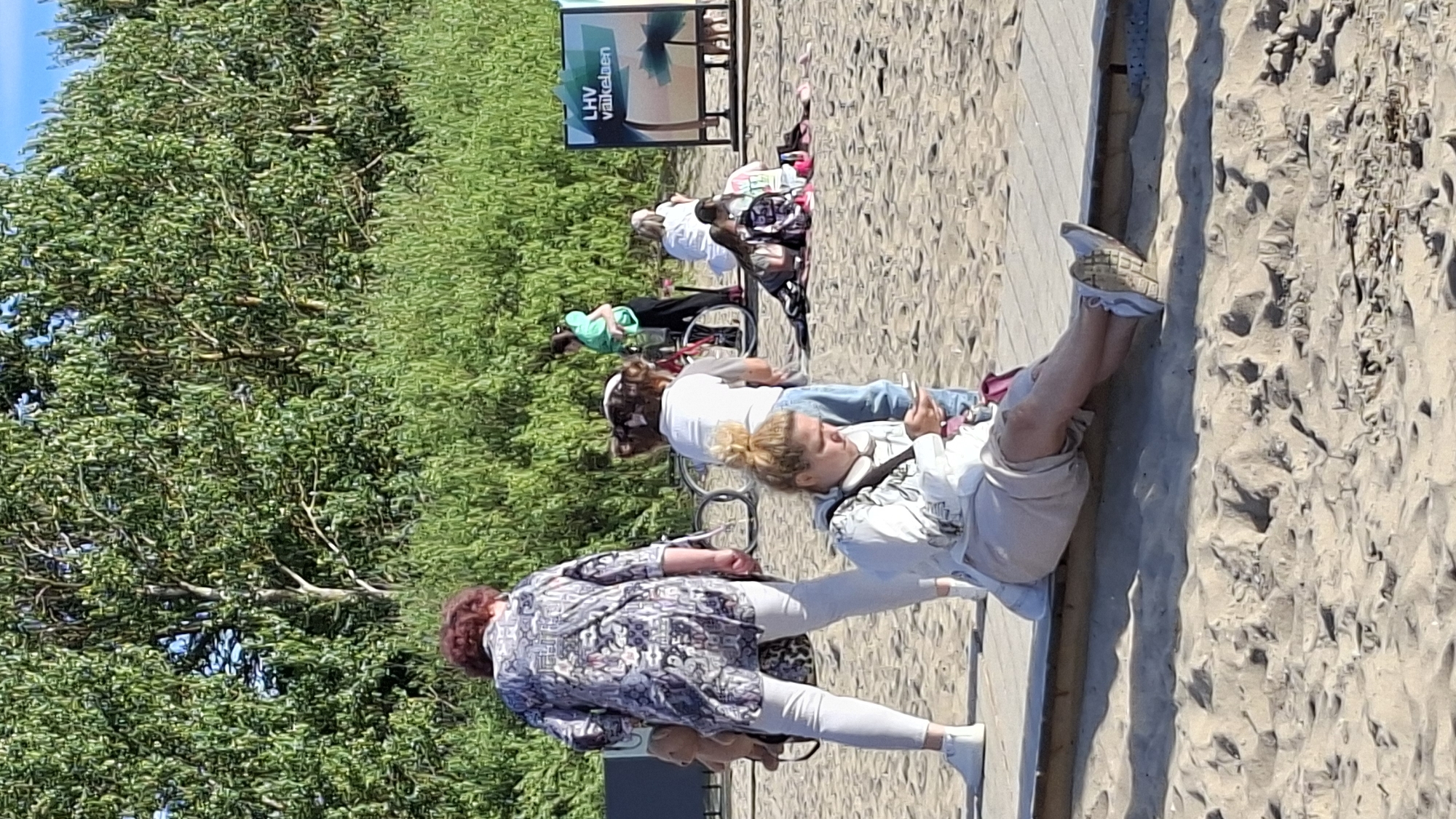 The image depicts a sunny outdoor scene at a beach in Tallinn, Estonia, likely Pirita beach due to the presence of a sign that says "LHV Väikelaps" which translates to "LHV Toddler". In the foreground, a woman with blonde hair is sitting on a wooden boardwalk, looking at her phone. She is wearing a light-colored hooded jacket and light-colored shorts. Behind her and to the left, a woman with dark, curly hair is standing. She is wearing a patterned blouse and white pants, with a stuffed teddy bear attached to her backpack. Further in the background, a group of people are gathered on the sand. One person is wearing a green shirt and black pants. Another person is wearing a white top. Several people are sitting on blankets. In the mid-ground, there are two bicycles. The environment includes a sandy beach, a wooden boardwalk, lush green vegetation, and a bright blue sky.