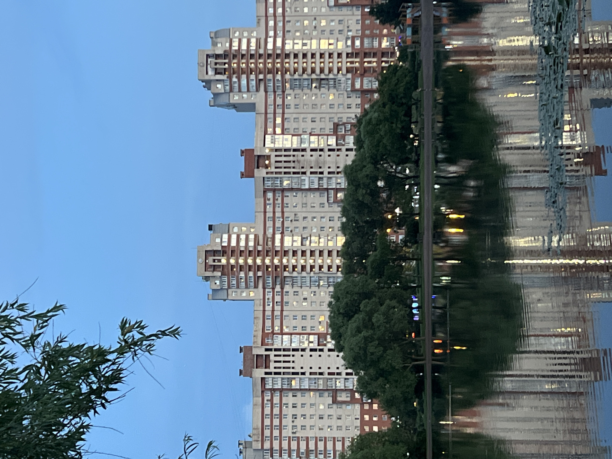 This is a vertical, outdoor, eye-level, medium shot photo taken in Saint Petersburg, Russia. The image captures a modern, multi-story building with a beige and red facade, reflecting in the calm water of a canal. The building is characterized by its numerous windows, some of which are illuminated, suggesting it is either evening or a cloudy day. The architecture features an unusual, stepped design on the left side of the building.
The reflection in the water is distorted by ripples, creating an abstract impression of the building, trees, and lights. Dark green trees line the edge of the canal between the building and the water, partially obscuring the lower floors of the building. In the foreground on the left, the branches of a leafy tree are visible against a clear blue sky. The overall impression is one of urban serenity, with the natural element of the water and trees contrasting with the man-made structure.
The time of day appears to be dusk or early evening, given the illuminated windows and the blue hue of the sky. No people are visible in the image. No text is visible. The scene evokes a sense of quiet contemplation or an appreciation for urban landscape photography.