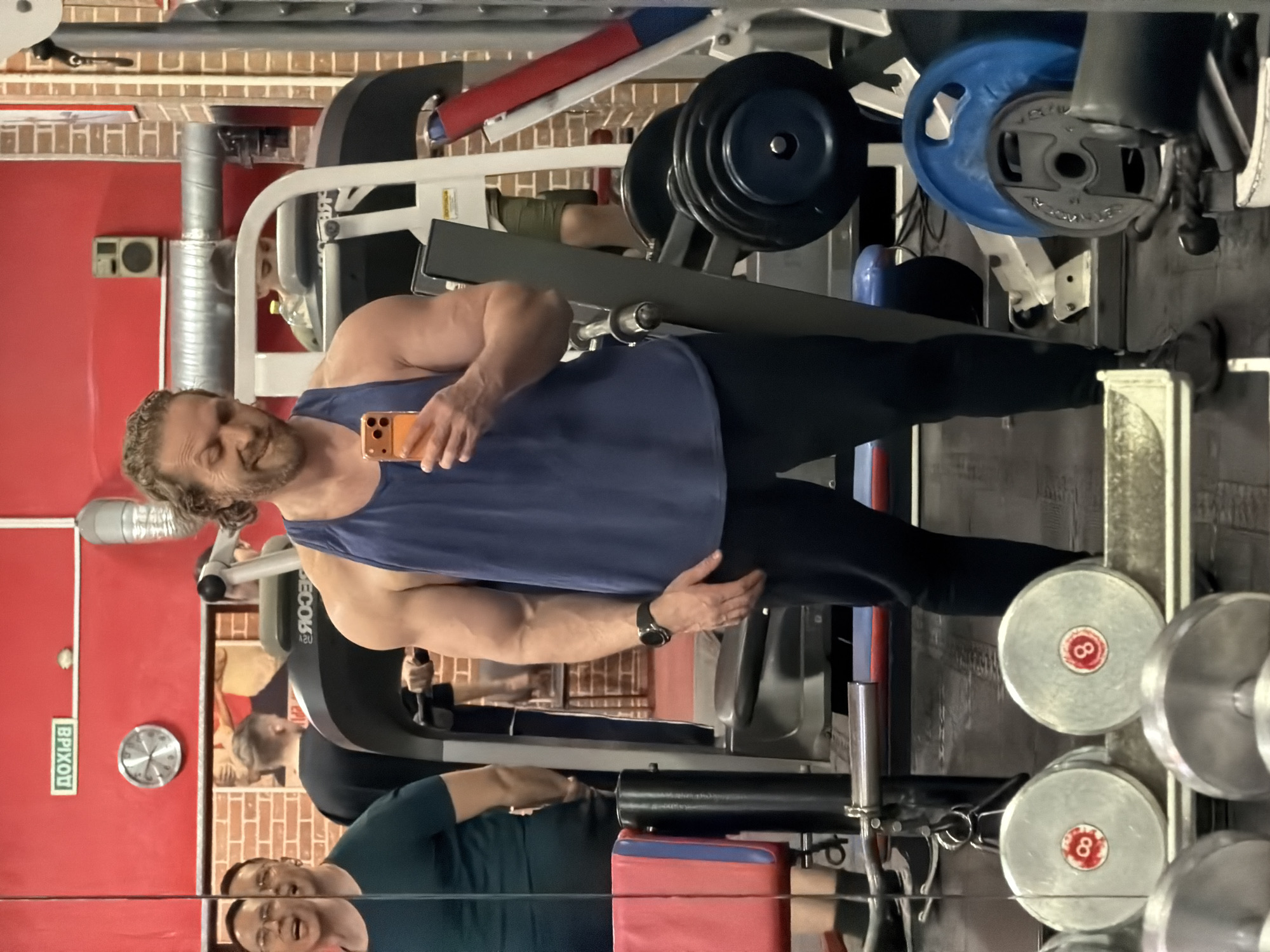 The image shows a man taking a selfie in a gym. He is wearing a blue tank top and black pants. In the background, there are weight machines, dumbbells, and a treadmill. The gym has red and brick walls. A Russian sign says "ВЫХОД," meaning "Exit" in Russian. The reflection in the mirror shows other people working out. Based on the sign and the architecture, the location is likely in Russia, possibly Saint Petersburg.