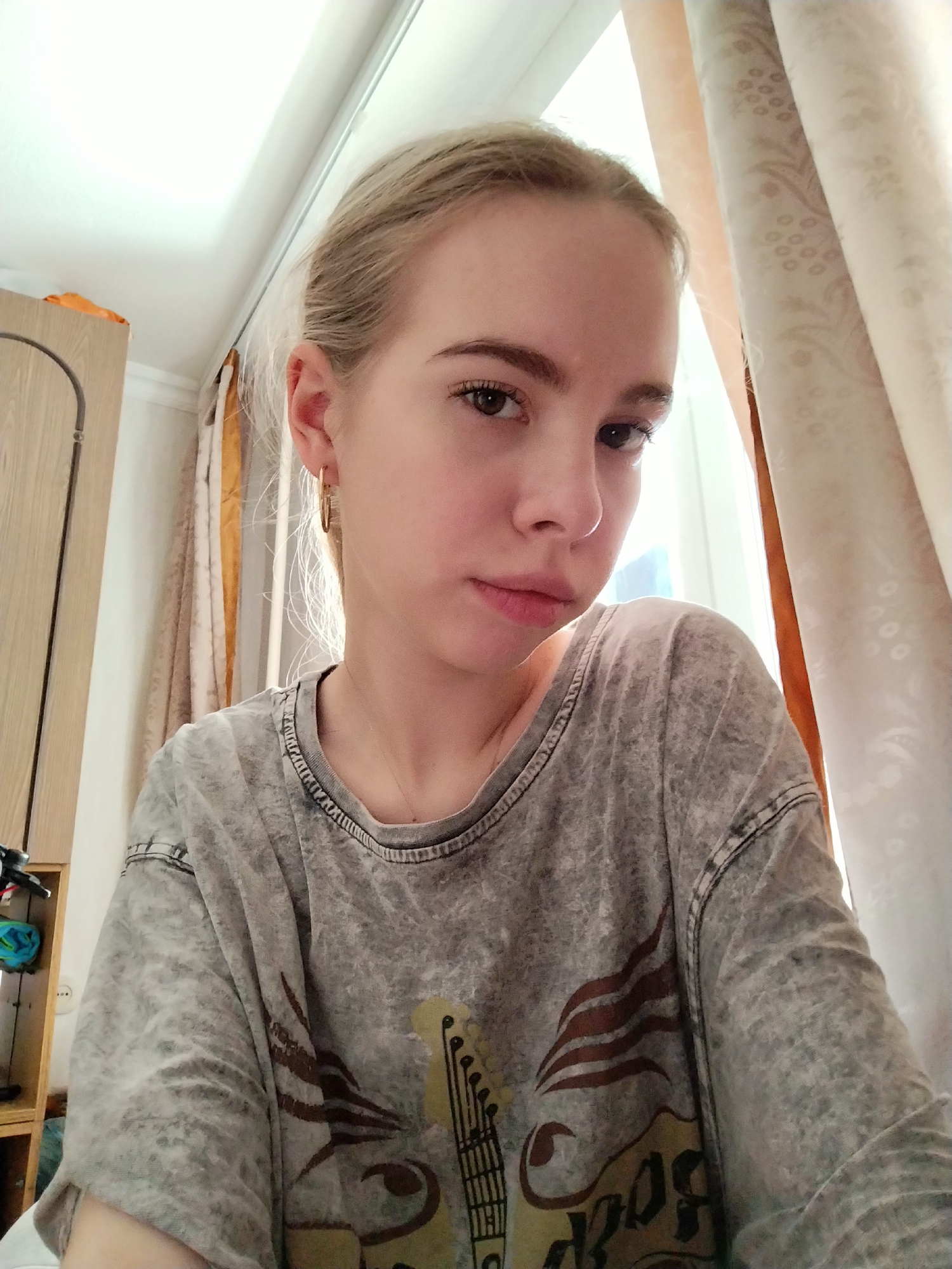 A young Caucasian woman with blonde hair pulled back poses for a selfie. She is wearing a light grey, acid-washed t-shirt with a graphic design of a guitar and flames, along with the word "Rock" visible. A gold hoop earring is visible in her left ear. The background shows a window with sheer, patterned curtains and a glimpse of what appears to be a wardrobe or cabinet to the left. The lighting suggests it is daytime, with natural light coming from the window. The woman's expression is neutral, with her head slightly tilted. The overall setting appears to be indoors, likely a bedroom or living space.