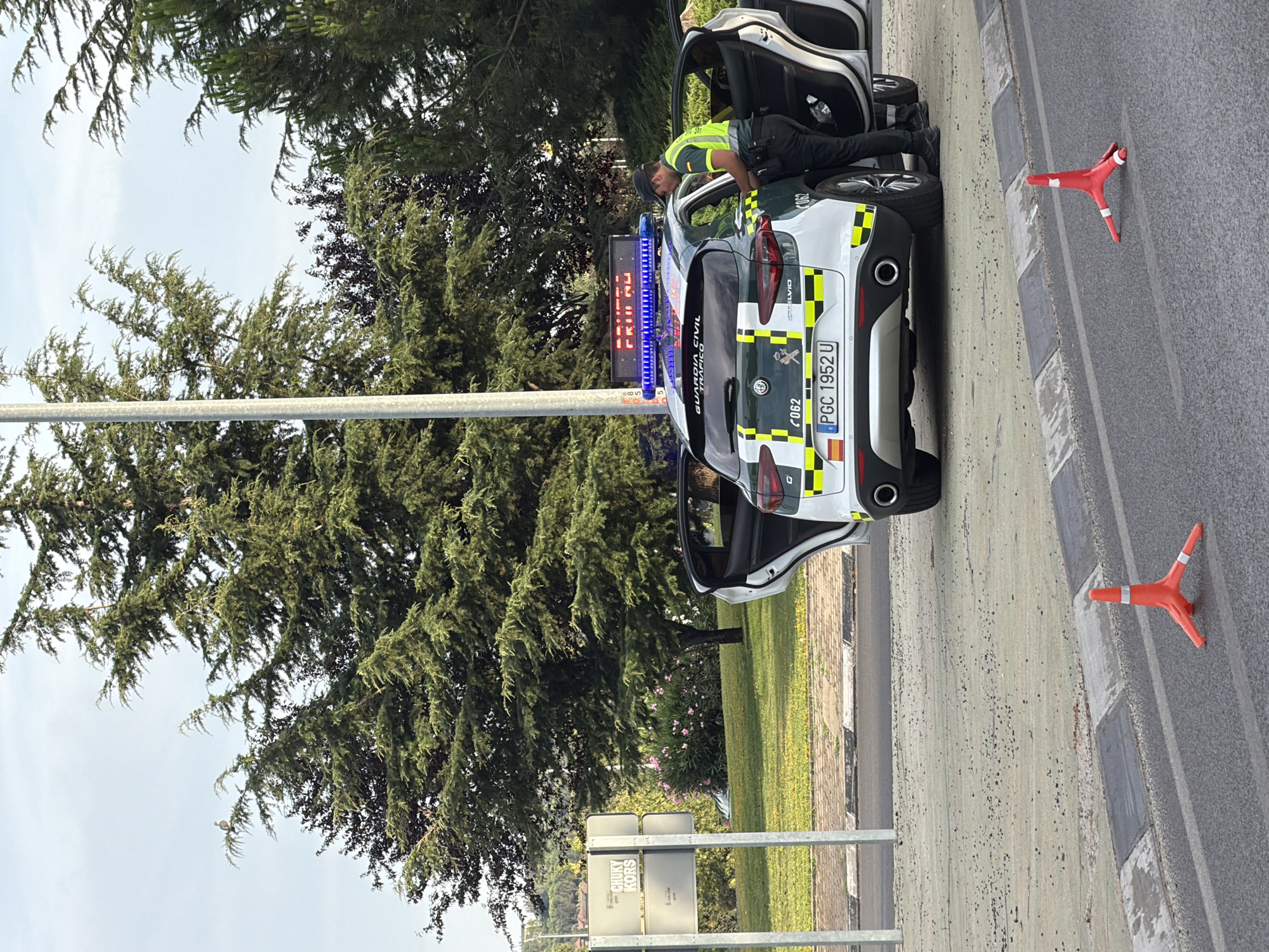 In Collado Villalba, Spain, a Guardia Civil traffic vehicle with its emergency lights activated is parked on the side of a road. A Guardia Civil officer wearing a high-visibility vest and a baseball cap is standing by the open driver's side door, appearing to interact with someone or something inside the vehicle. The vehicle has "GUARDIA CIVIL TRAFICO" written on the rear and a license plate reading "PGC 1952 U". A digital sign above the vehicle displays red text, possibly a warning or informational message. Two orange traffic cones are placed on the road in the foreground. The scene takes place outdoors, with trees and greenery visible in the background, suggesting a suburban or park-like setting.