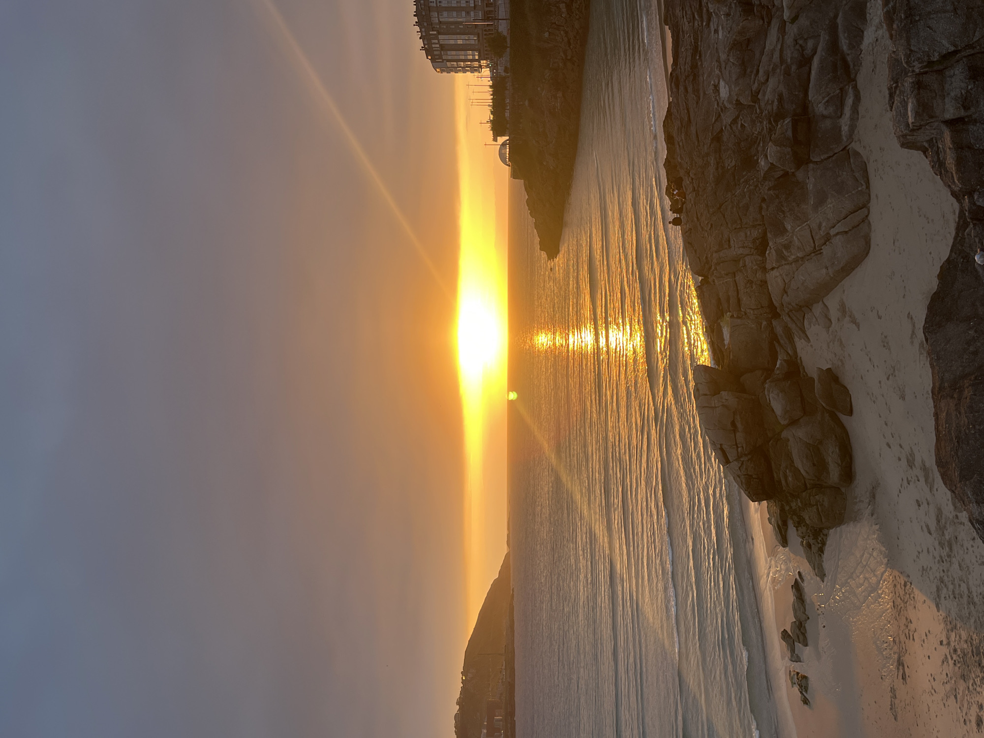 The image depicts a sunset scene at a beach in A Coruña, Spain. The sun is setting over the ocean, casting a strong golden light that reflects on the water's surface. Buildings and cliff edges are visible along the coastline on either side of the frame, with some structures appearing in silhouette. There are visible beachgoers amongst the rocks.