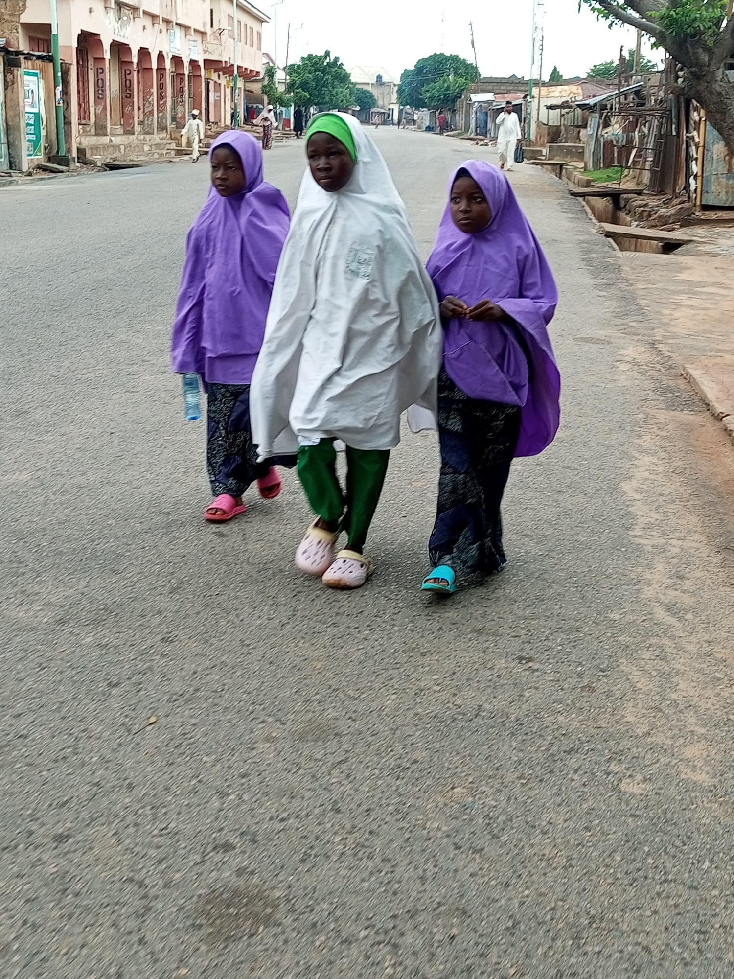 Three young girls wearing hijabs are walking on a paved street in Gombe, Nigeria. The girl in the center wears a white hijab and green pants. The girl to her left wears a purple hijab and dark patterned pants, and carries a blue water bottle. The girl to her right also wears a purple hijab and dark patterned pants. They are walking away from the camera. In the background, buildings line the street, and a few other people are visible walking. The street appears to be in a commercial or residential area.
