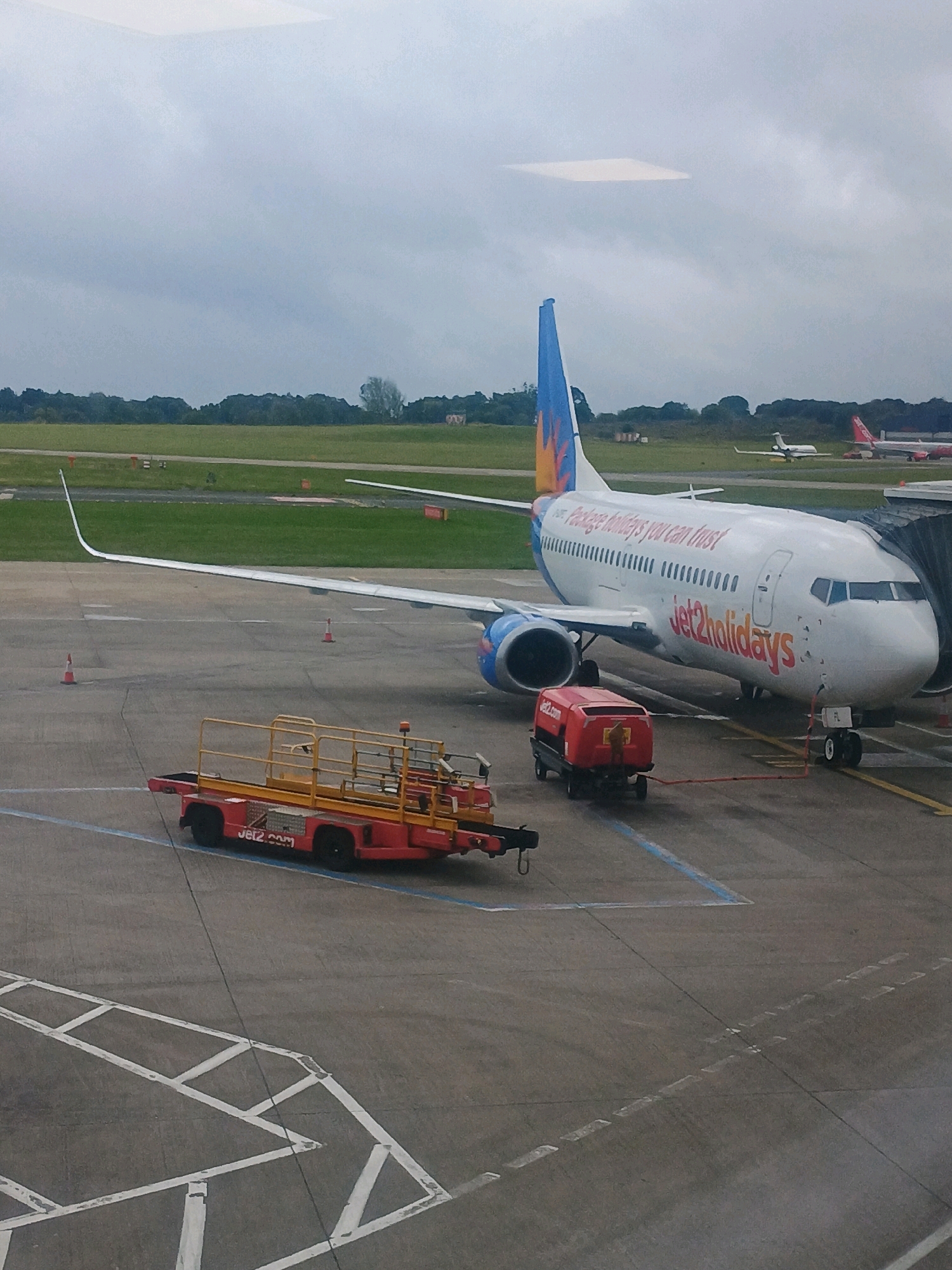 An aerial view captures an airport tarmac under a cloudy sky. A Jet2holidays airplane, white with orange and blue accents, is parked at a gate, connected to a jet bridge. The words "Jet2holidays" are prominently displayed on the fuselage. In front of the plane, a red baggage tug with a yellow rack is parked. Smaller orange traffic cones are scattered on the tarmac. In the background, more airplanes are visible, along with grassy fields and trees. The overall scene depicts a typical airport environment on a cloudy day.