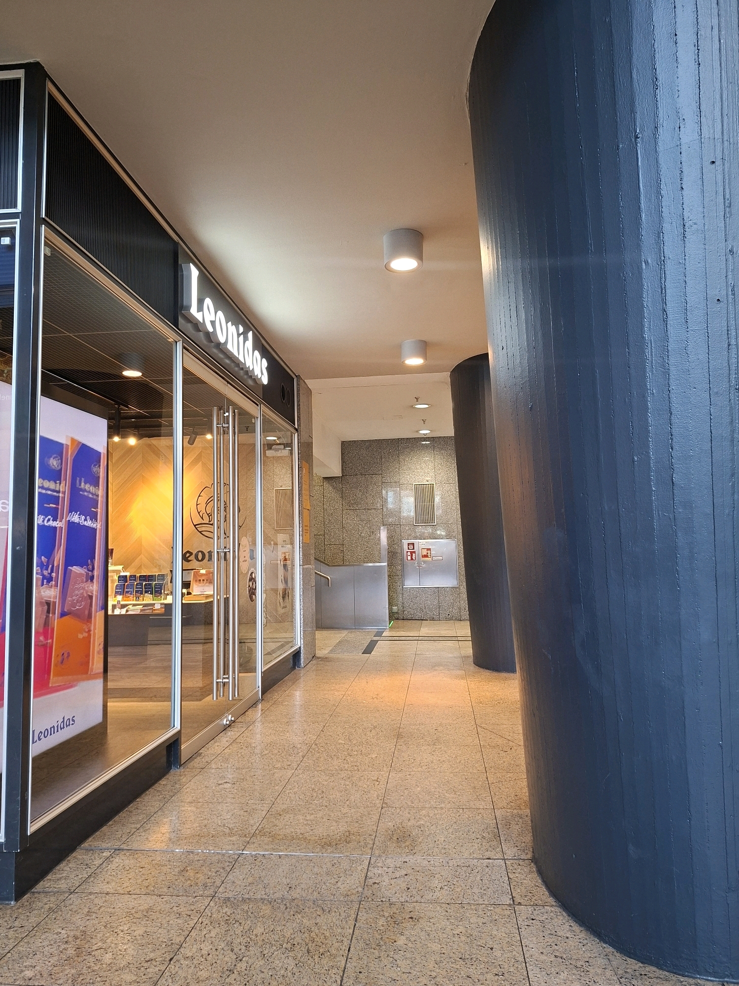 The image shows the entrance to a Leonidas chocolate shop in Cologne, Germany. The shop has a glass facade with double doors. Inside, shelves display chocolates and promotional materials. The exterior features a large, dark blue, cylindrical pillar. The floor is tiled with a speckled pattern, and the ceiling has recessed lighting. In the background, there is a grey tiled wall with a metal fire alarm box and a fire extinguisher. The overall scene depicts a retail environment in an urban setting.