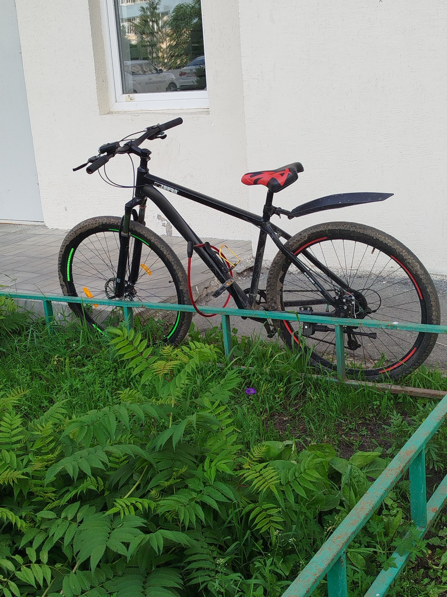 A black bicycle with a red and black seat is parked next to a white building. The bicycle has a U-lock attached to the frame and a bottle cage. The front tire has green accents, and the rear tire has red accents. The bicycle is parked on a paved surface next to a patch of green grass and some foliage. A green metal fence is in the foreground. The building has a window with a reflection of cars and trees. This scene is located on a street in Yekaterinburg, Russia.