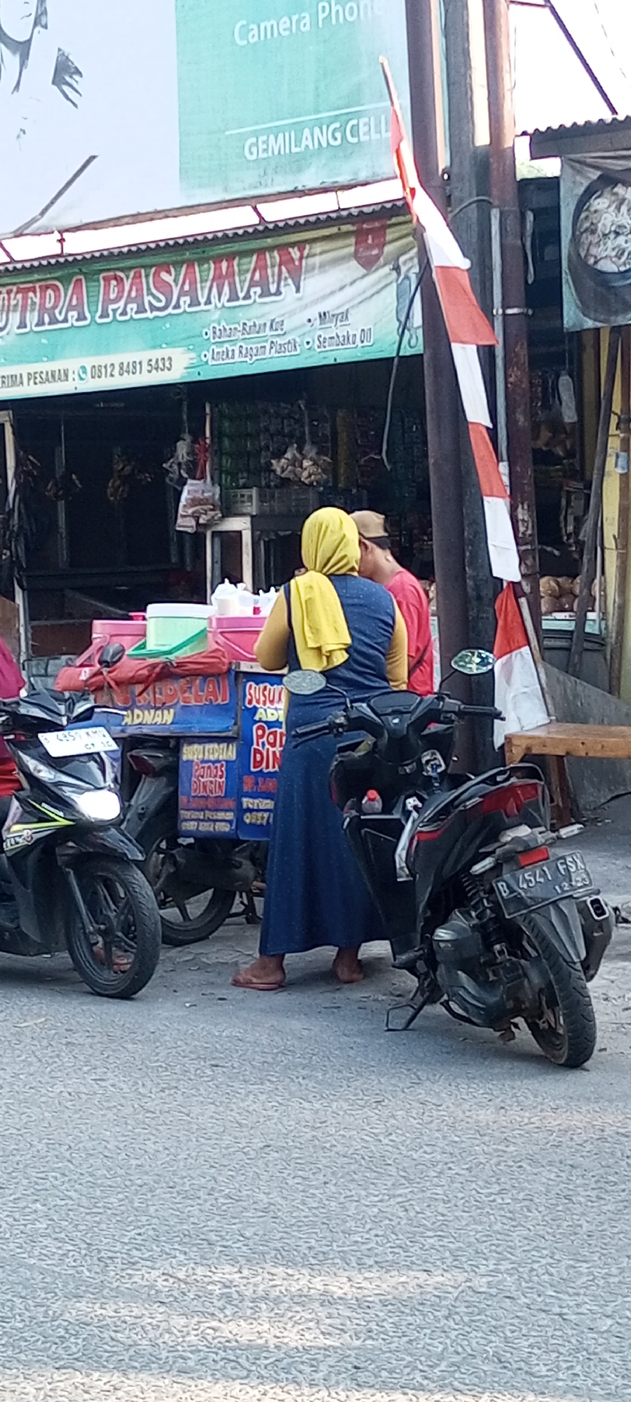 In Setu, Indonesia, on a paved road with a textured surface, a street-side vendor stall is visible, primarily selling food items. Two individuals are interacting at the stall, a woman wearing a yellow hijab and a dark blue dress, and a person with a tan cap and a red shirt standing behind her. The woman is facing away from the camera, towards the stall.
The stall itself is decorated with a banner that reads "PUTRA PASAMAN" in large, red and green lettering. Below this, smaller text indicates items sold: "Bahan-Bahan Kue" (Baking Ingredients), "Minyak" (Oil), "Aneka Ragam Plastik" (Various Plastics), and "Sembako DO" (DO Groceries). Adjacent to this banner, a blue board displays more text, including "SUSU" (Milk), "AD" (Advertisement or Additive), "PAR" (likely part of a word like 'Pari' for specific food item or 'Pare' for bitter melon, or could be a brand name), and "DIN" (likely part of a word like 'Dinding' meaning wall or 'Dinding' used as a container term). Further text on this blue board includes "SUSU KEDELAI" (Soy Milk), "PANGS" (likely "Pangsit" meaning wonton or dumpling), and "DINRIN" (likely part of "Didingin" meaning chilled or frozen). There are also price markings visible.
Two motorcycles are parked on either side of the stall. The motorcycle on the left is black with white and green accents, and its headlights are on. The license plate is visible, reading "B 4357 KMU" with smaller text below. The motorcycle on the right is also black, and its license plate reads "B 4541 FSN", with "12-23" indicating its expiration date.
In the background, a larger banner is visible, with text that includes "Camera Phone" and "GEMILANG CELL". A red and white Indonesian flag is also prominently displayed, attached to a pole. The overall scene suggests a typical street vendor operation in a bustling area, offering a variety of goods and services.