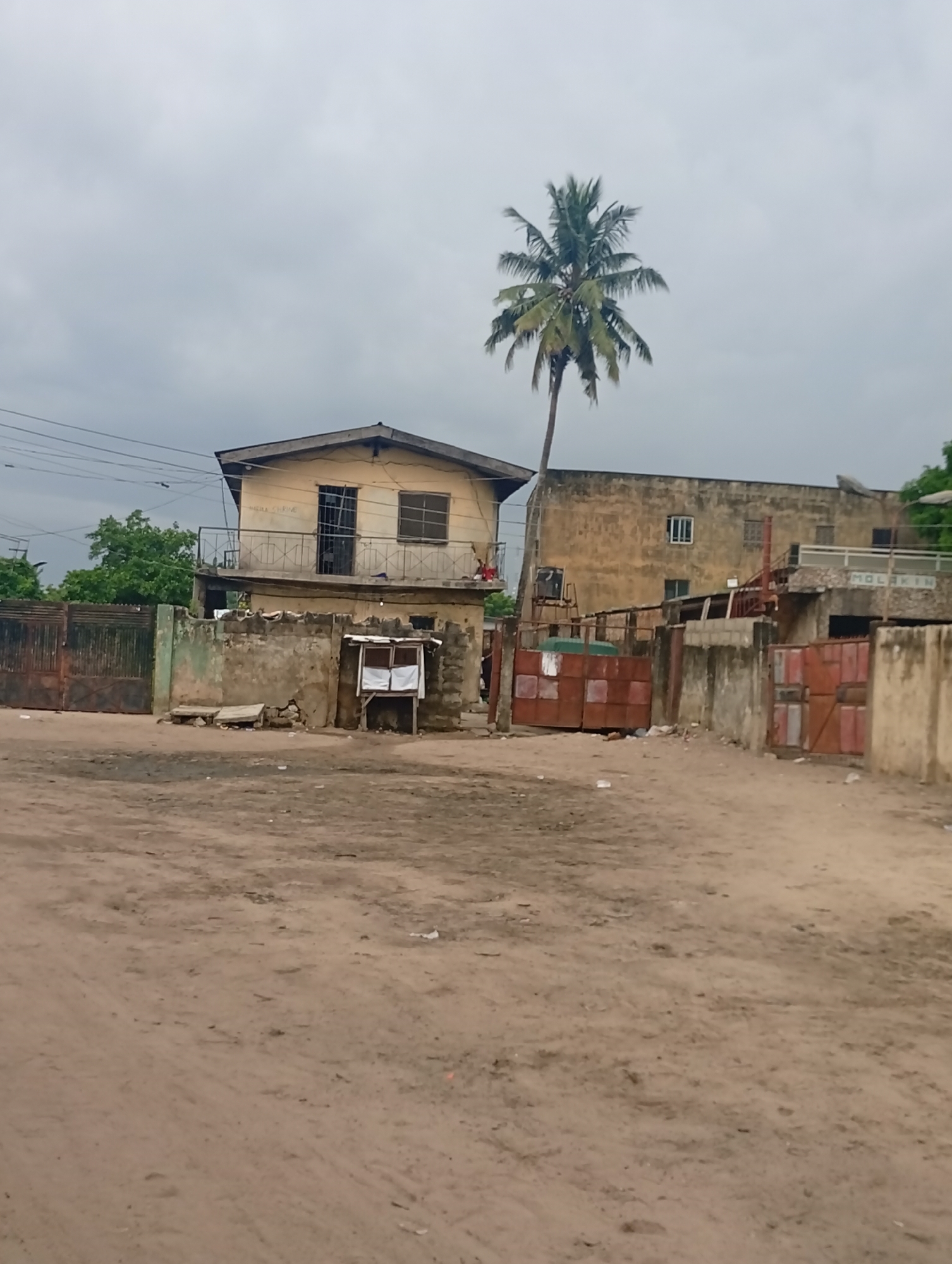 The media file presents an outdoor scene in Ijanikin, Nigeria, under an overcast sky, suggesting a cloudy day without direct sunlight. The foreground features a wide, unpaved dirt ground, marked with tire tracks and some debris.
In the midground, several structures are visible. On the left, a two-story, light yellow house with a gabled roof stands behind a low concrete wall and a large, partially rusted metal gate. The house has a balcony on the upper floor with a metal railing. Faint, possibly faded text "MEWA CHRINE" can be discerned on the wall above a lower window on the balcony level. In front of this house, a small, makeshift stand or kiosk with white signs stands next to the concrete wall; the text on these signs is illegible.
To the right, a tall palm tree with a dense canopy of fronds rises prominently, towering over the buildings. Further right, a larger, light brown concrete building with multiple windows extends across the frame. This building also sits behind a concrete wall and rusted metal gates, and features an external staircase. The word "MOLAKIN" is visibly painted on a section of its exterior wall. Power lines are strung across the upper left portion of the image.
The scene depicts a static street or open area in what appears to be a residential or mixed-use neighborhood. No people or explicit activities are taking place. The overall setting reflects a functional, lived-in environment with practical, unornamented architecture.