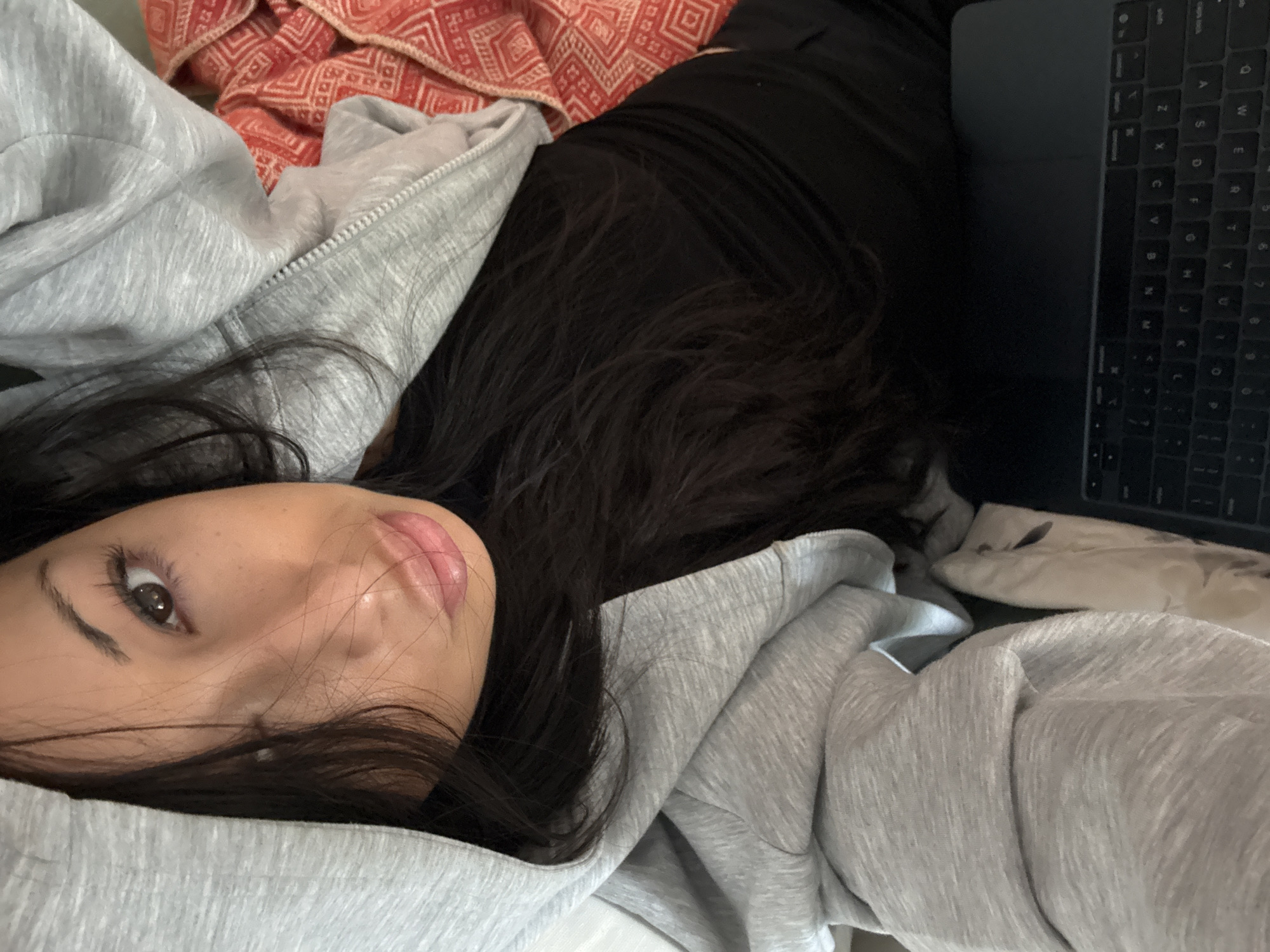 A close-up, angled shot captures a person lying down, possibly in bed. The individual has dark hair and is wearing a light gray hooded sweatshirt. A laptop, with its keyboard visible, rests beside them. The background includes a glimpse of a patterned red and white textile. The setting appears to be indoors, likely a private residence.