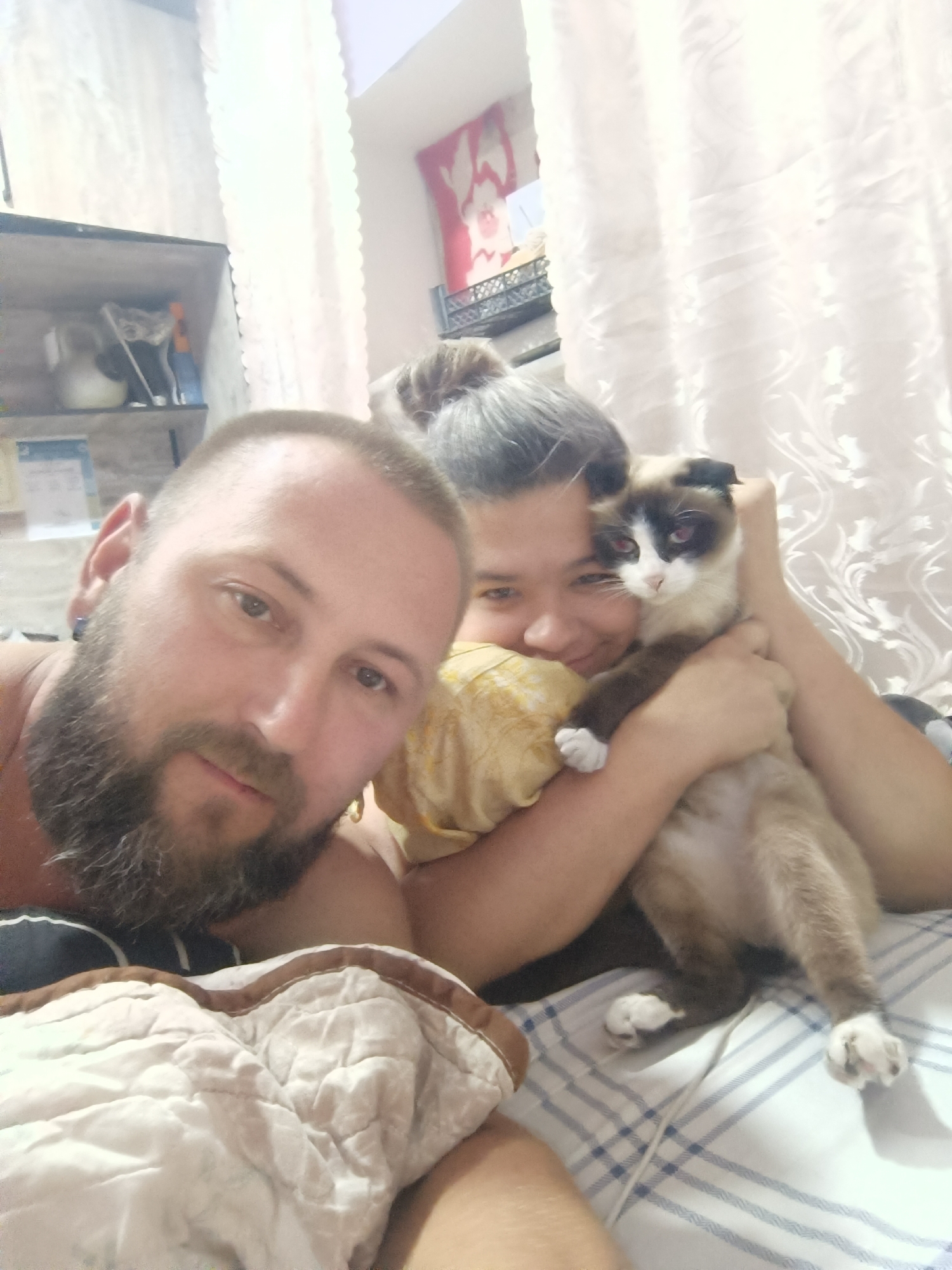 In a dimly lit room, a man with a beard and a woman are lying down, with a cat on the woman's lap. The man is in the foreground, looking at the camera. The woman is behind him, holding the cat. The room appears to be a bedroom, with a bed, curtains, and shelves in the background. The scene does not contain any discernible landmarks or indicators specific to Taraz city administration, Kazakhstan.