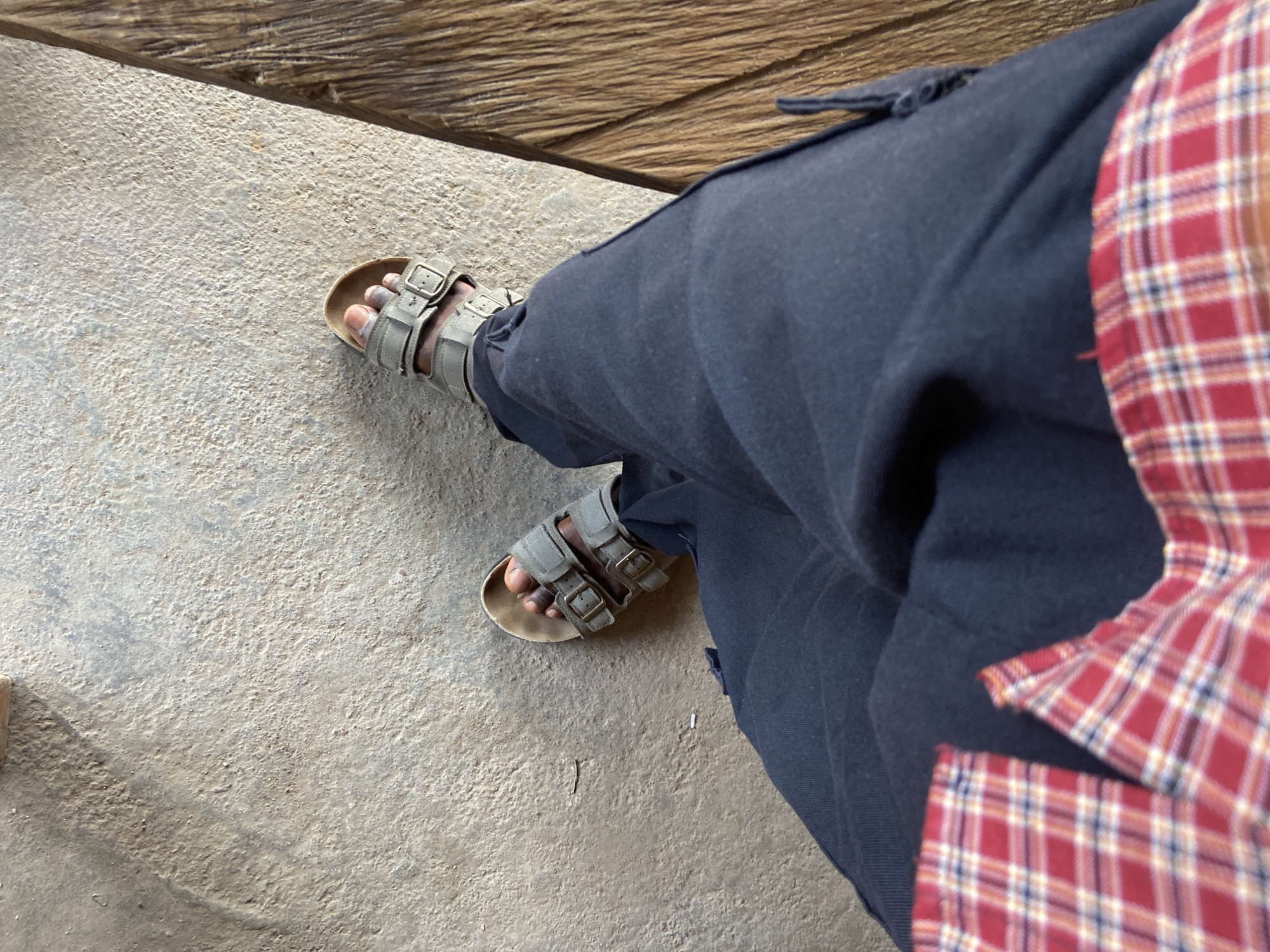 An overhead shot captures a person's lower leg and foot, positioned on a rough, light-colored concrete or cement ground in Mararaba, Nigeria. The person is wearing a pair of light olive-green or grey sandals with two adjustable buckled straps and a visible footbed, resembling a Birkenstock style. Their skin tone appears to be dark, and some whitish residue, possibly dust or dried material, is present on their toes and foot.
The person is dressed in dark blue trousers or cargo pants, and a red and white plaid shirt is visible on the right side of the frame, partially obscured. A distinct wooden surface, possibly a bench, step, or beam, runs diagonally across the top of the image, showcasing its natural grain and texture. The lighting indicates it is daytime, with shadows suggesting direct light, but no specific weather conditions are discernible. The overall perspective is from above, looking down towards the ground and the person's leg. There is no visible text in the image.