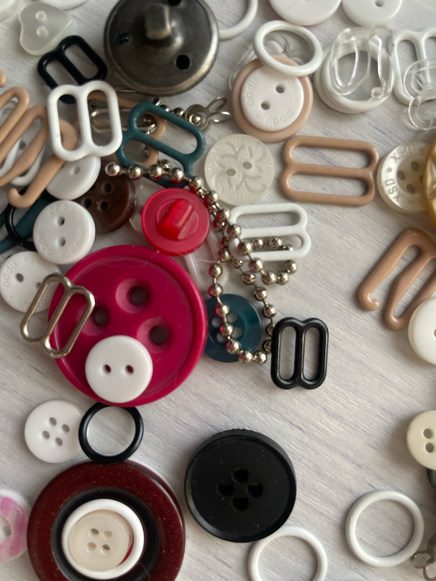 The image displays a collection of sewing notions, predominantly buttons and strap adjusters, arranged on a light-colored, textured surface. The items include various sizes, colors, and materials such as plastic, metal, and possibly ceramic. There are no people or animals visible in the image, and no discernible elements suggest a specific location within the city of Atarfe, Spain. The context is purely that of crafting supplies.