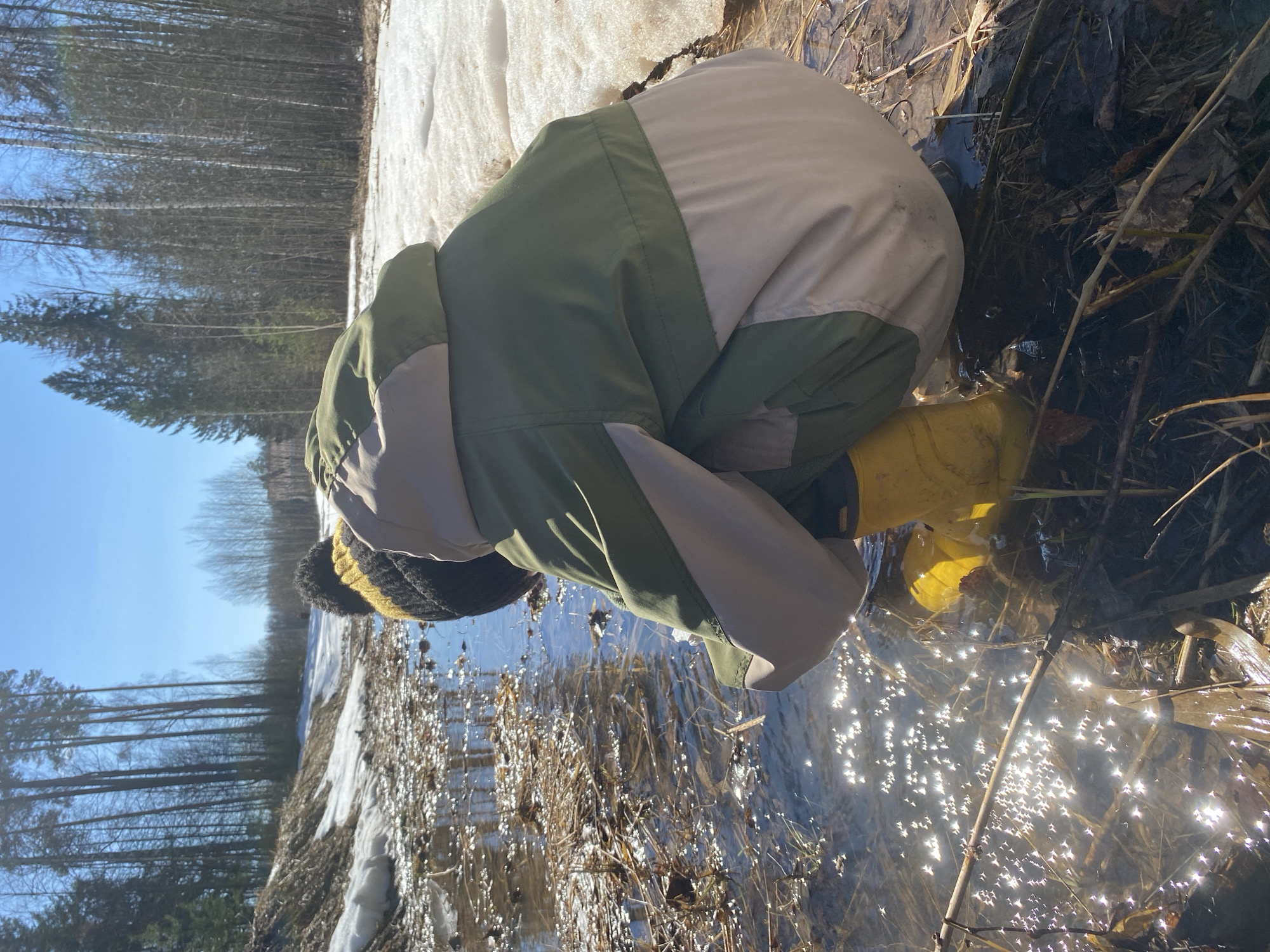 A child wearing a green, beige, and black coat, yellow boots, and a black and yellow hat is squatting at the edge of a partially melted snow bank next to a pool of water. The water is reflecting the bright sunlight. On the other side of the melting snow bank are bare trees. The location appears to be a rural area near Noviy, Russia.