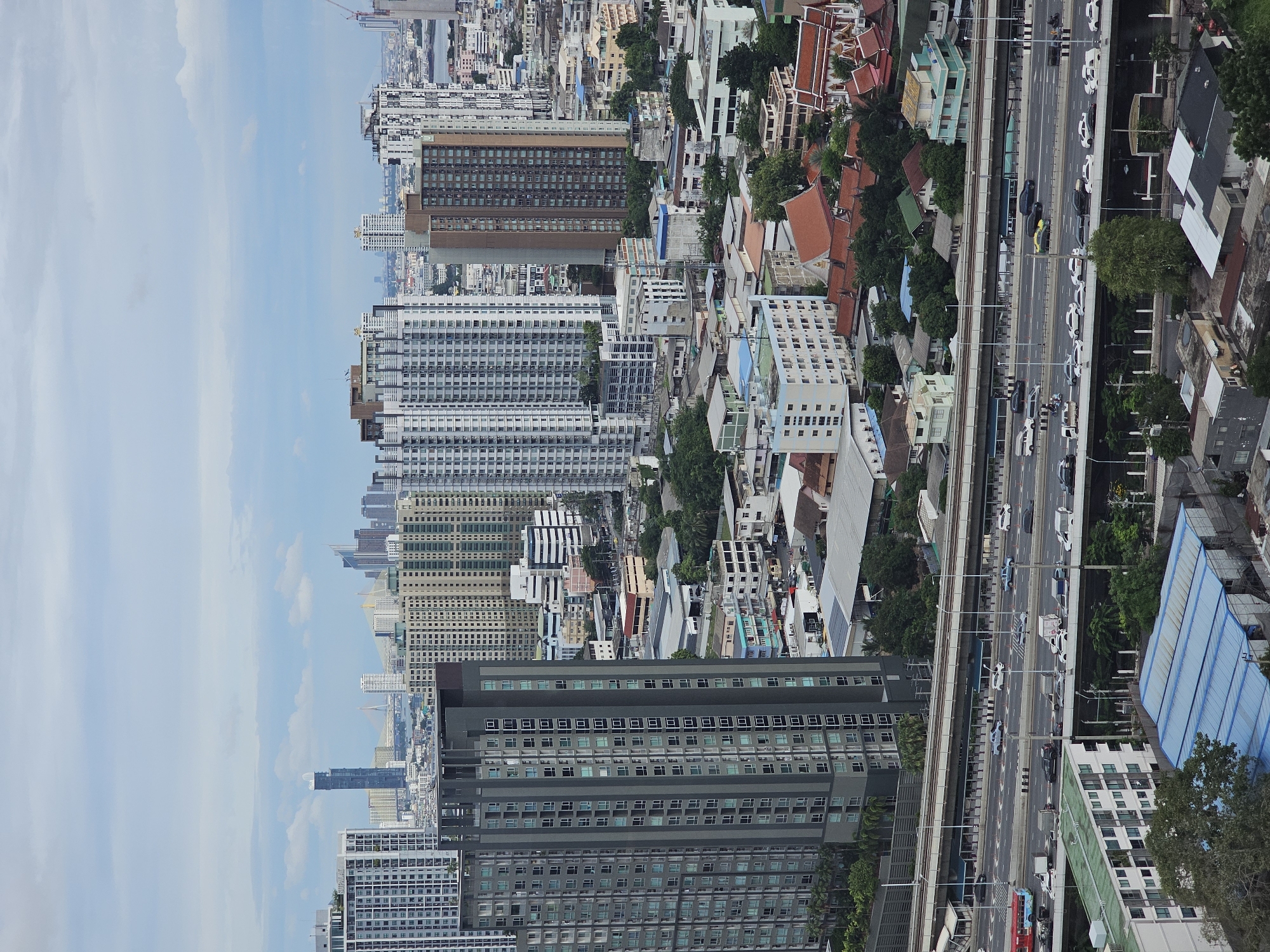This is a high-angle aerial view of a dense urban landscape in Bangkok, Thailand, dominated by numerous high-rise buildings and a multi-level highway. In the foreground, a grey high-rise apartment building with a grid-like pattern of windows is prominent. Adjacent to it, a multi-lane highway is visible, with cars, trucks, and a red bus in transit. The highway is elevated and passes over a lower-level area with more residential buildings and some industrial-looking structures with blue roofs. Further in the background, the cityscape extends with a variety of apartment blocks, some with light-colored facades and others with darker tones. The sky is overcast with scattered clouds. The architecture suggests a modern Asian metropolis, with a mix of residential and commercial structures, and the presence of a major transportation artery indicates a busy, developed area. The general scene conveys the bustling nature of a large city.
