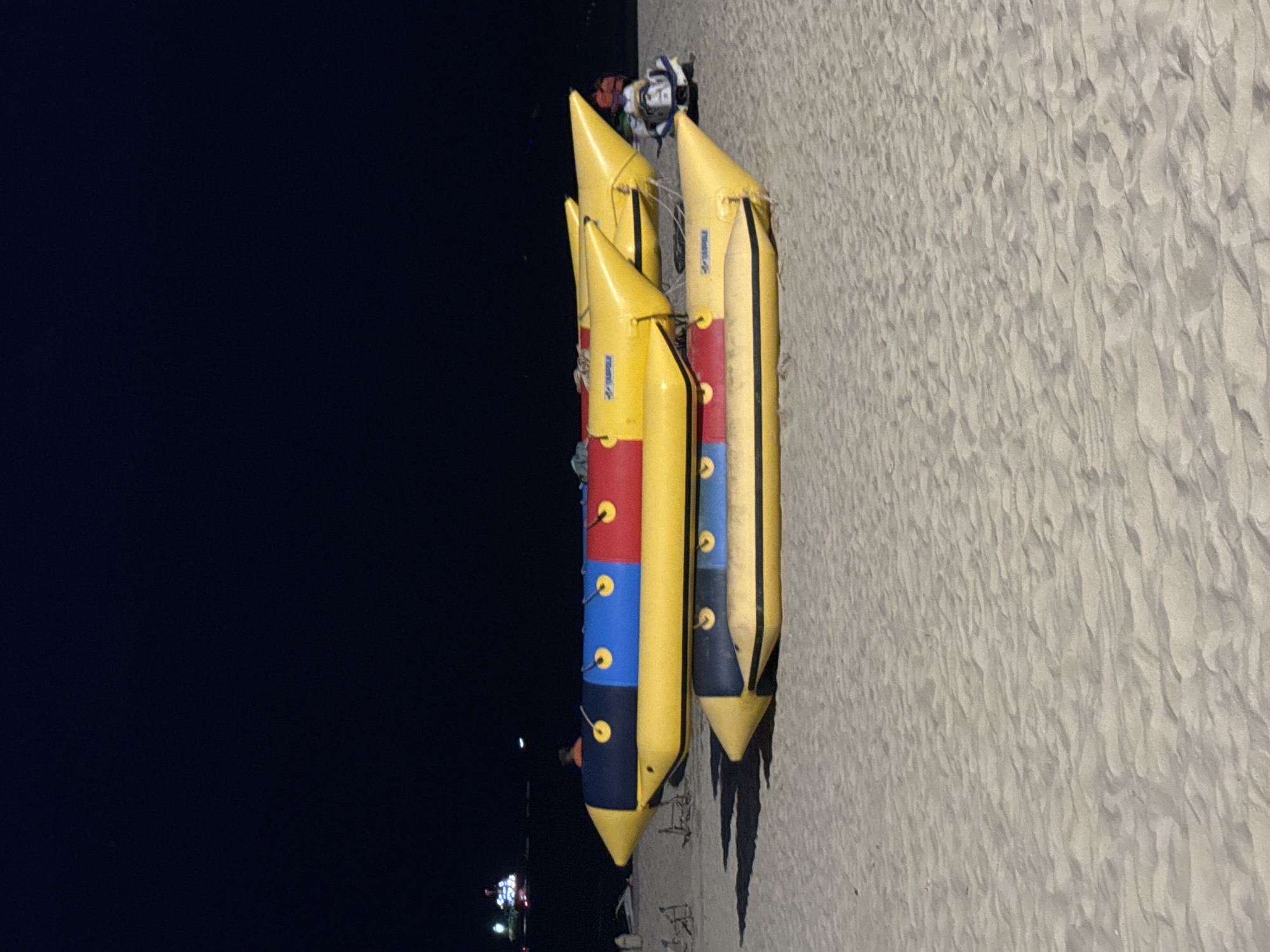 Two inflatable banana boats are parked on a sandy beach at night. The top boat is yellow with blue and red sections and yellow handles. The bottom boat is mostly yellow with a black stripe and yellow handles. In the background, the dark ocean is visible with some lights from a distant structure or boat. The sky is completely dark. The beach is covered in sand.
