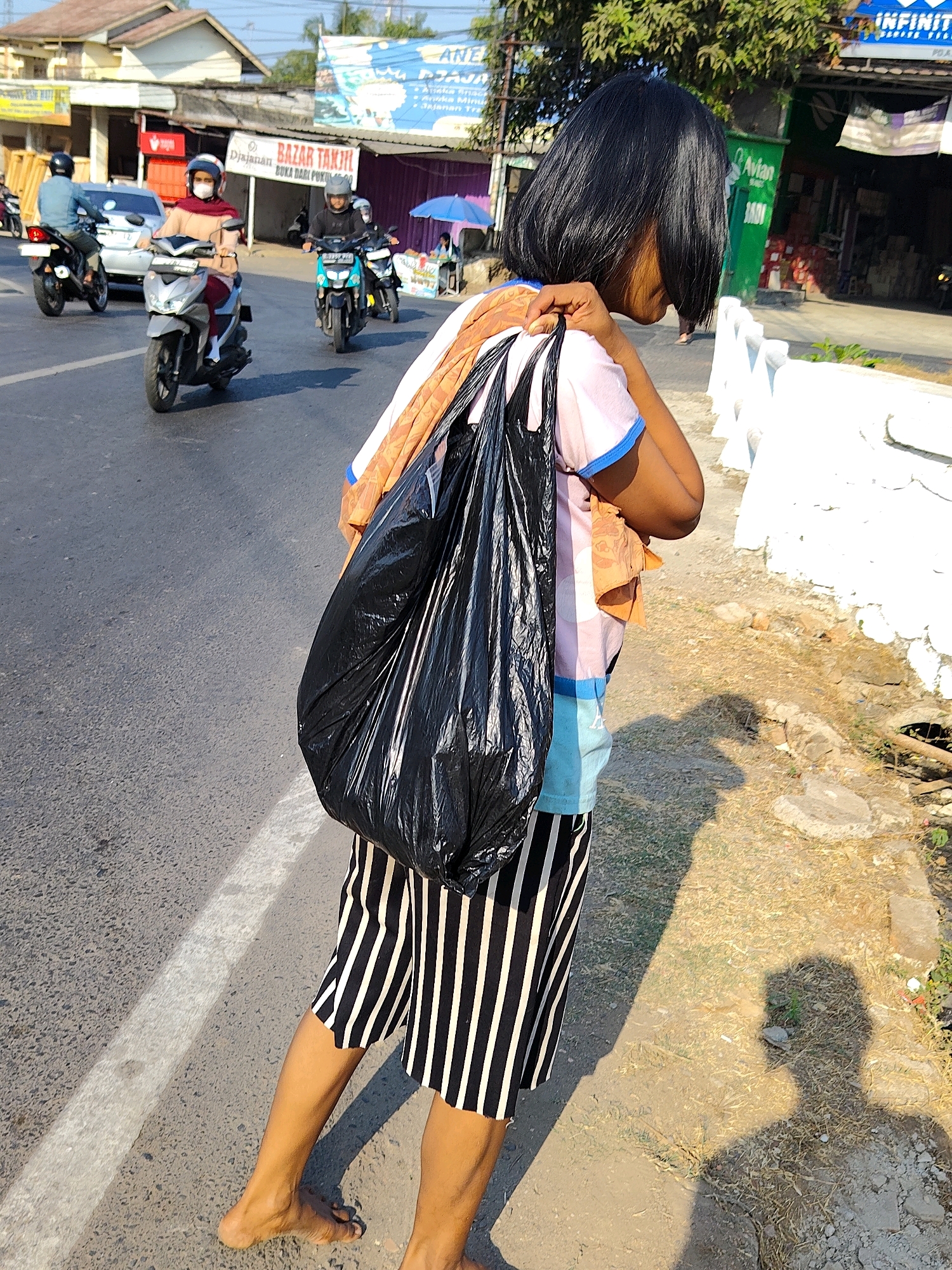 A young female, seen from the right side, walks barefoot on the side of a road in Purwakarta, Indonesia, carrying a large black plastic bag over her right shoulder. She wears a light-colored t-shirt with blue trim and striped shorts. A small orange cloth is tucked into the waistband of her shorts. In the background, several motorcycles are visible on the road, with riders wearing helmets. Buildings and trees line the street. The ground beside the road is dry and covered with sparse vegetation and debris. Sunlight casts a shadow of the girl on the ground.