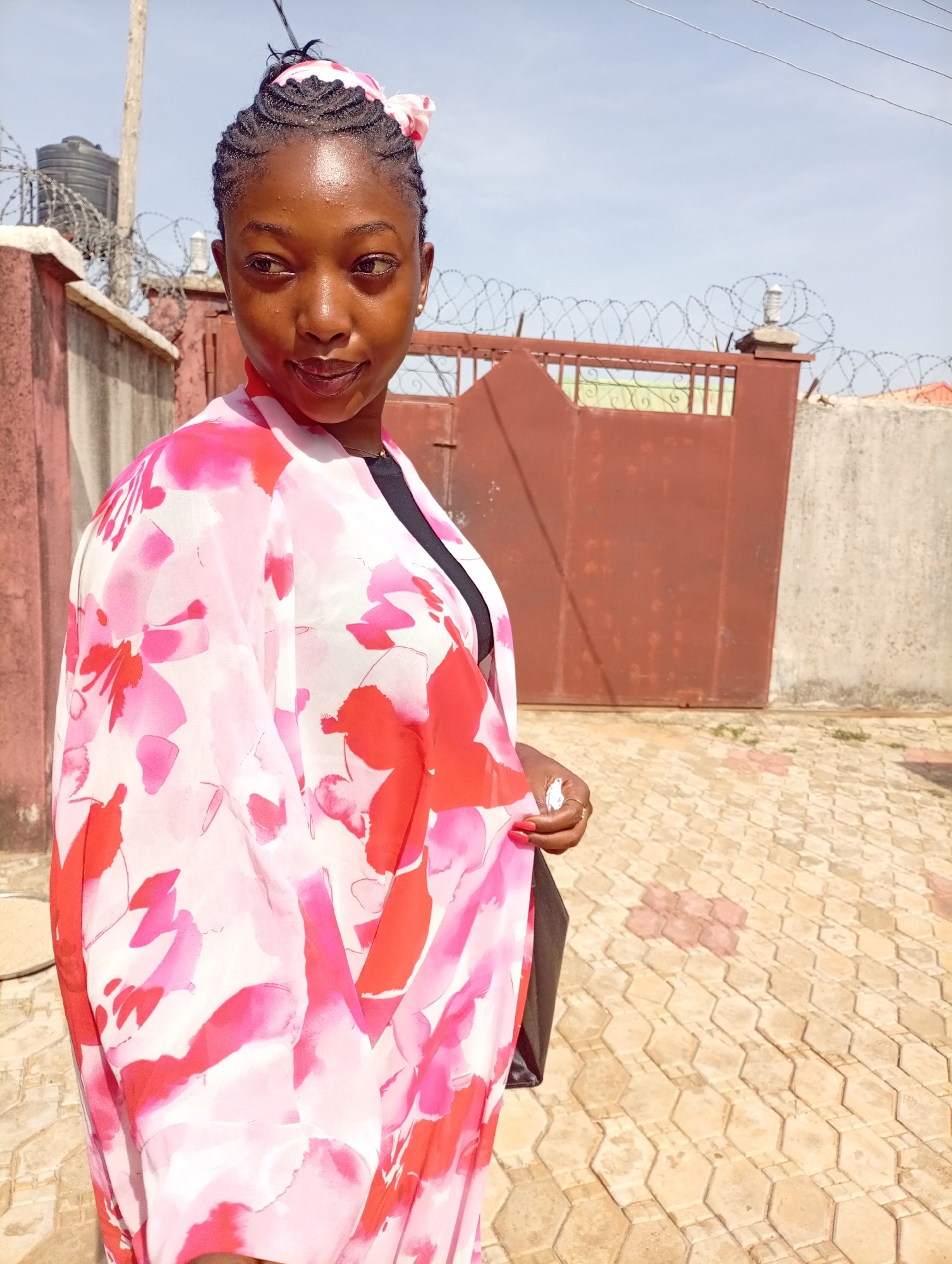 A young African woman is standing outdoors on a patterned paved area. She is wearing a floral kimono-style garment over a dark top. The woman has braided hair adorned with a pink floral hairband. Behind her is a wall with a red metal gate topped with barbed wire. A second, taller wall is visible to the right of the gate. The location is the city of Unknown, Unknown.