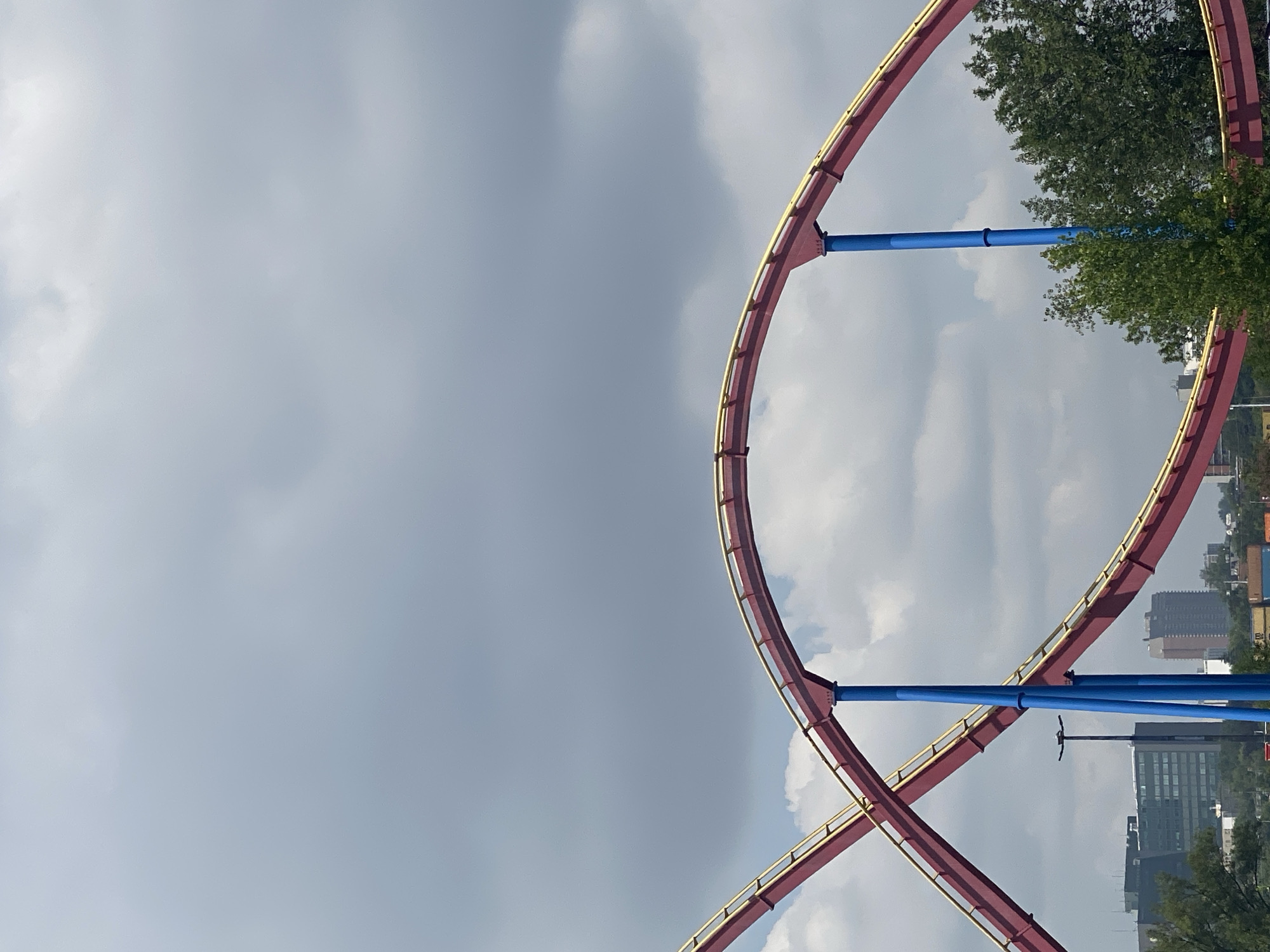 This is a low-angle, eye-level, full shot of a roller coaster track at an amusement park, likely La Ronde, located on Île Sainte-Hélène in Montreal, Canada. The roller coaster track, painted red with yellow trim, forms two large curves, one ascending and one descending, supported by bright blue vertical pillars. In the background, a partly cloudy sky with grey and white formations dominates the upper portion of the frame. Below the roller coaster, a city skyline is visible, featuring several buildings of varying heights and architectural styles, including a modern glass-fronted skyscraper. Trees with lush green foliage are scattered throughout the scene, adding a touch of nature to the urban landscape. A streetlamp with a single light is visible to the left of the roller coaster track. The overall scene is static, with no people or explicit action taking place. The lighting suggests it is daytime, possibly late afternoon, given the light and shadow play on the clouds.