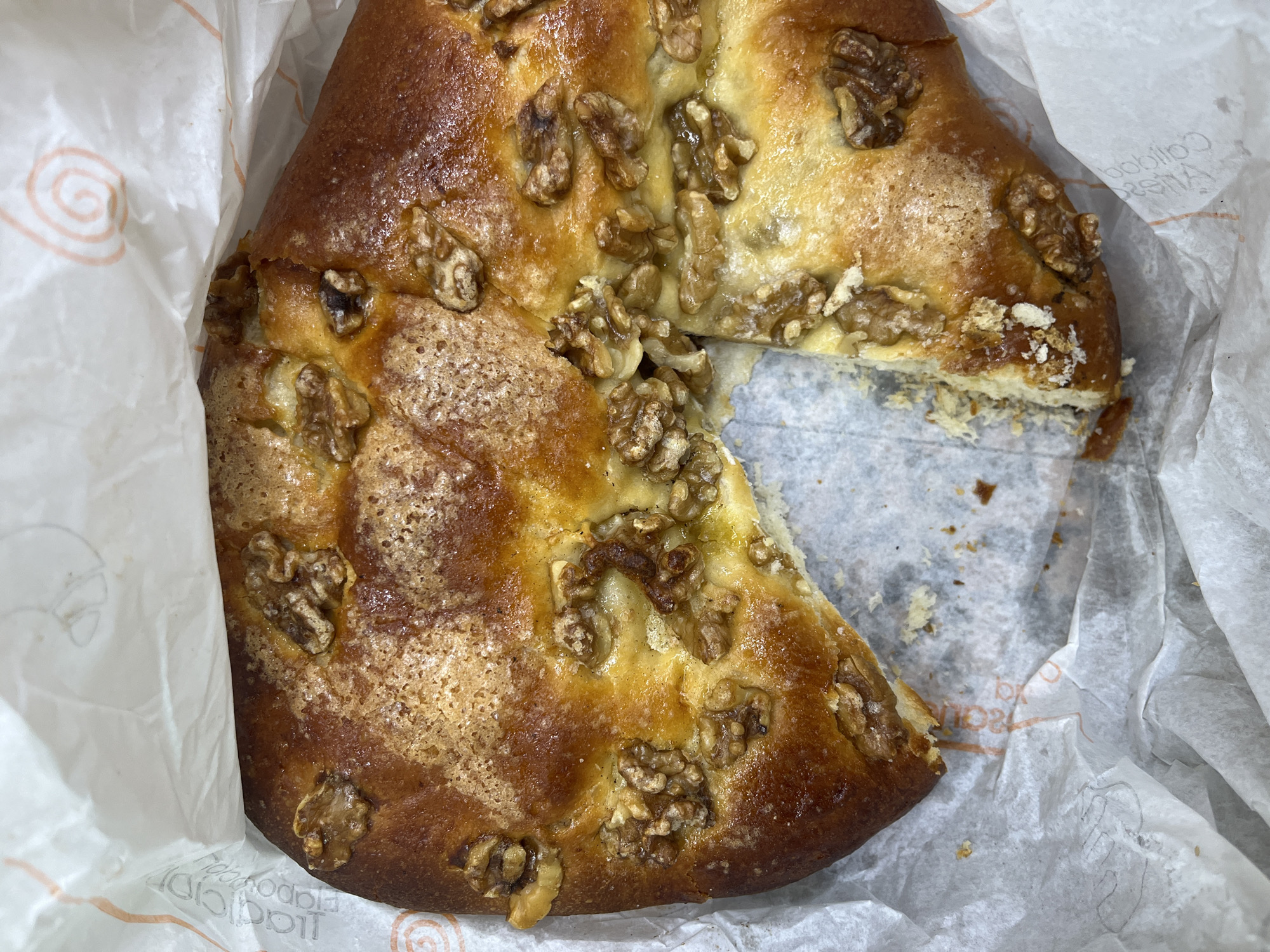 A round, golden-brown baked good, likely a sweet bread or cake, is presented within crinkled white paper packaging. The item is topped generously with numerous whole and halved walnuts, which are embedded in a glossy, slightly sticky glaze. The surface of the baked good shows variations in browning, with some areas appearing darker and more caramelized, particularly around the walnuts, and other areas lighter with a dusting that might be sugar crystals.
A triangular slice has been removed from the baked good, revealing its lighter, moist interior texture. Crumbs and smaller pieces of the pastry and walnuts are visible around the cut edge and on the paper lining.
The white paper wrapping is translucent in places and features orange-colored text and a spiral logo. Prominently visible in reverse on the paper are the words "Calidad Artesana" (Artisanal Quality), suggesting a handcrafted product. Further inverted text, which appears to read "Pollas," is also visible on the paper, potentially indicating the name of the bakery or producer.
The overall scene suggests a freshly prepared or purchased pastry, partially consumed, presented in its original packaging from a local establishment in Torrent, Spain.