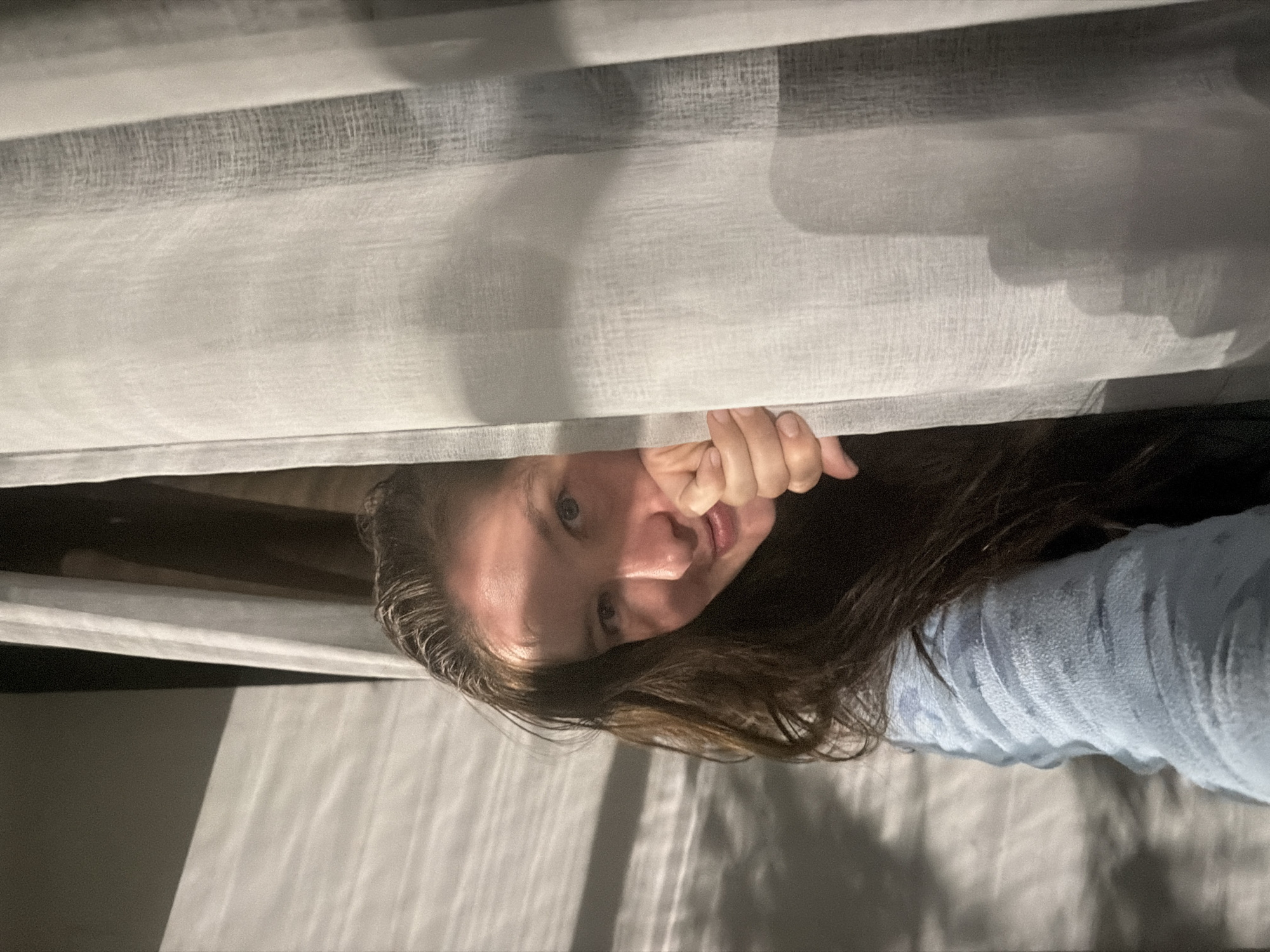A fair-skinned female with long, dark hair is looking through a gap between white or light gray curtains. Her right hand is visible near her face, partially obscuring her cheek. She is wearing a light blue garment. The environment consists of what appears to be interior lighting and the material of the curtains. The location, Staraya, Russia, cannot be determined from the image content.