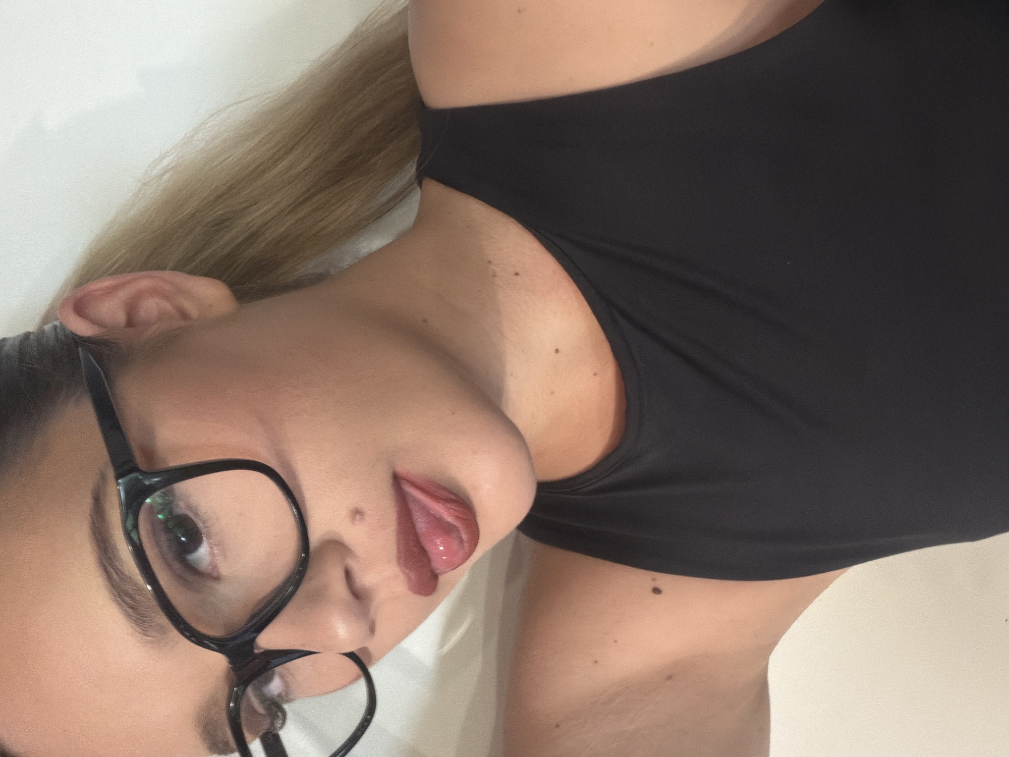 A young woman with blonde hair tied in a ponytail and wearing black-rimmed glasses is shown from the chest up. She is wearing a black tank top. Her tongue is sticking out. The background is a plain white wall. The image appears to be a selfie taken indoors.