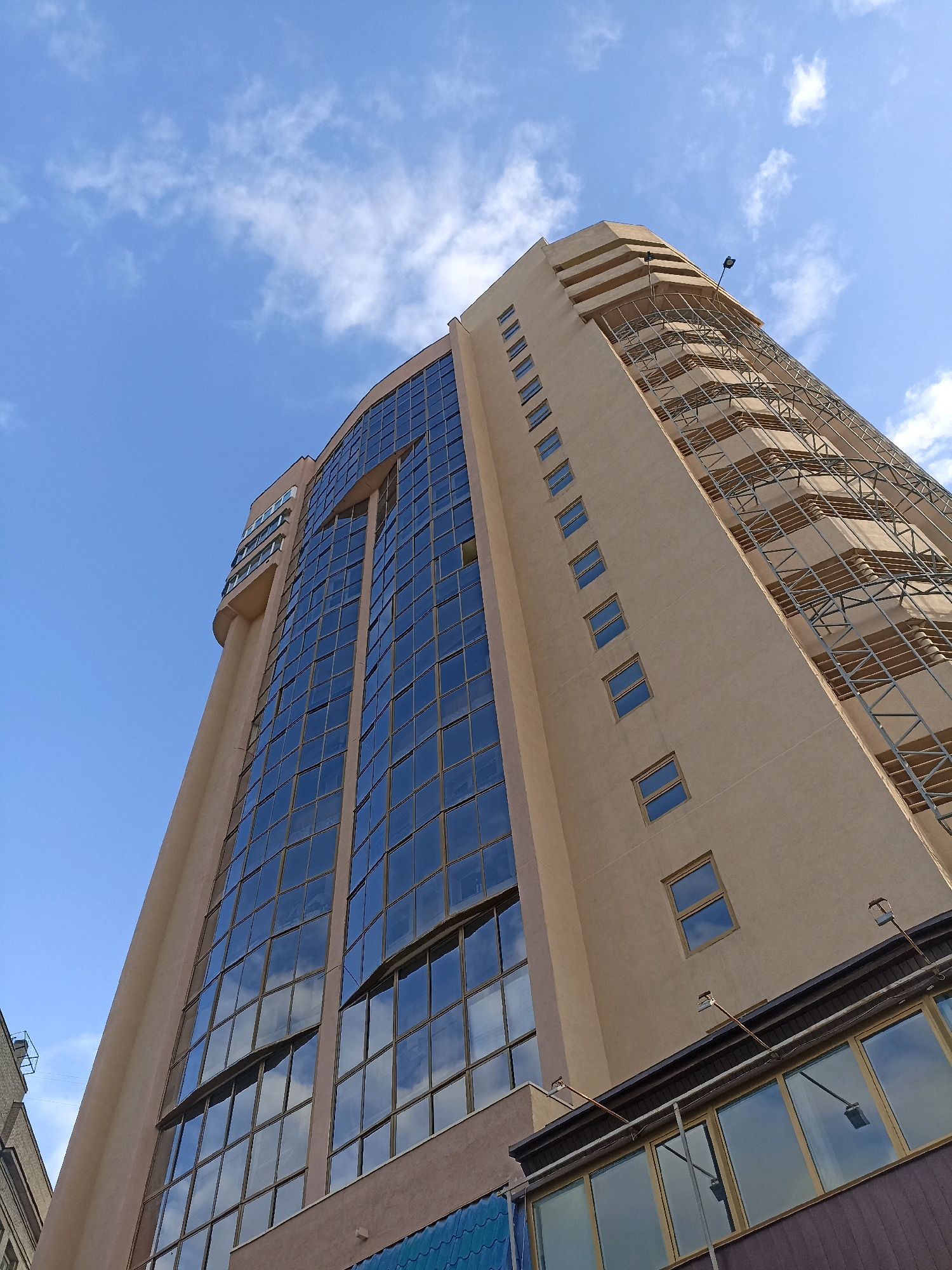 This is a low-angle, upward-facing, eye-level shot of a modern, multi-story building in Novosibirsk, Russia, under a bright blue sky with scattered white clouds. The building features a beige facade with a large, curved section dominated by a grid of reflective blue glass windows. Some of these windows are open. To the right of the glass facade, the building has a more angular design with a series of small, rectangular windows arranged vertically. A scaffolding-like structure, possibly for maintenance or construction, is visible along this section of the building. The top of the building has tiered, stepped sections and a visible light fixture. In the foreground, a lower structure with a dark frame and several large, horizontal windows can be seen. The overall impression is one of urban architecture.