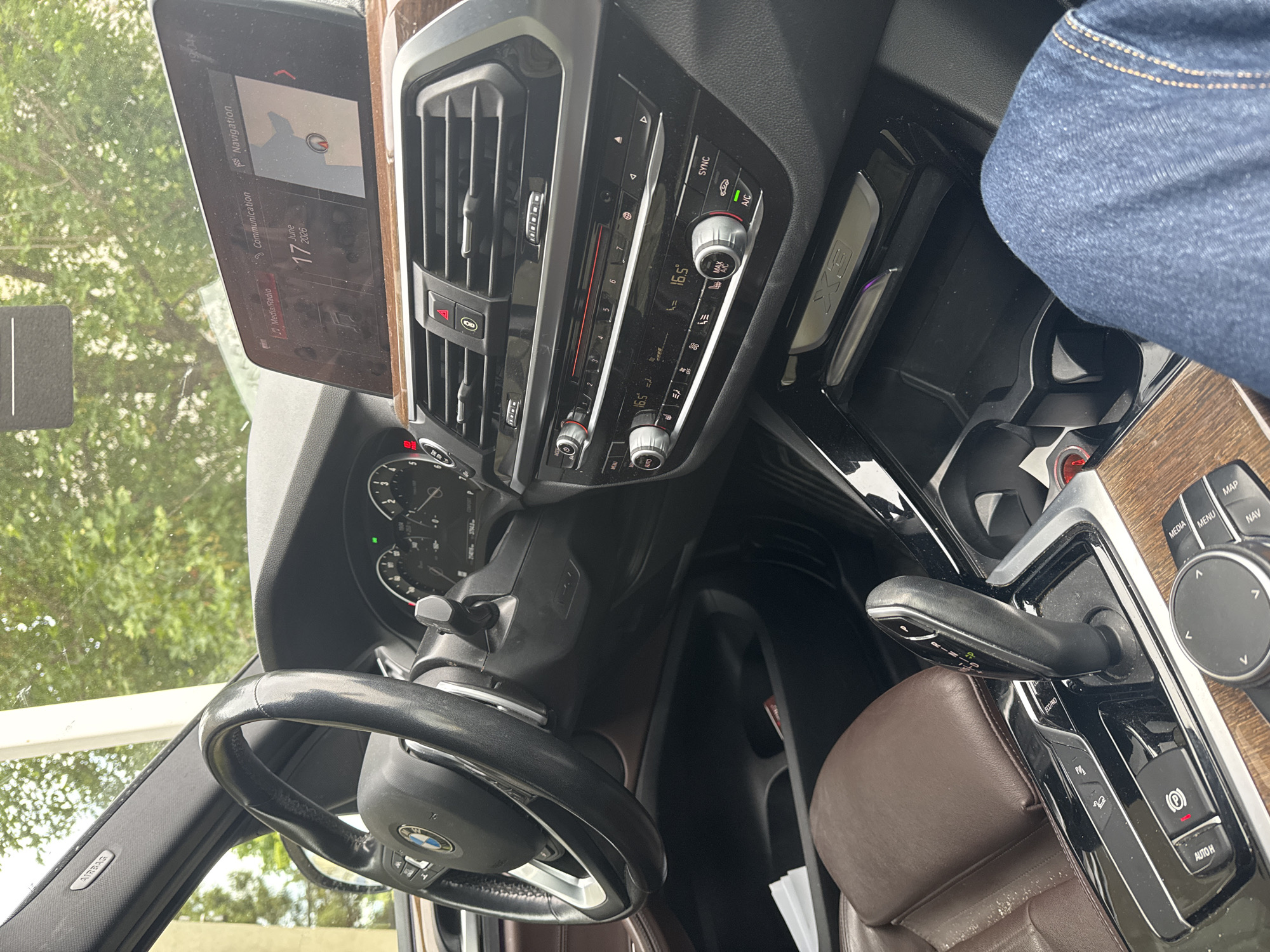 An interior, eye-level shot captures the dashboard of a BMW X3 parked in Nesebar, Bulgaria, under overcast skies. The leather-wrapped steering wheel, with a BMW logo at its center, dominates the left side of the frame. To the right, a prominent infotainment screen displays navigation information, time (19:04), and the date (June 17, 2026). Below the screen are air vents and climate controls, including temperature readouts of 16.5 degrees Celsius. A leather-trimmed gear shifter is visible in the foreground, with buttons for electronic parking brake and auto-hold. A person's legs, clad in blue jeans, are partially visible on the right. The interior features wood-grain trim accents. Outside, green foliage and trees are visible through the windshield.