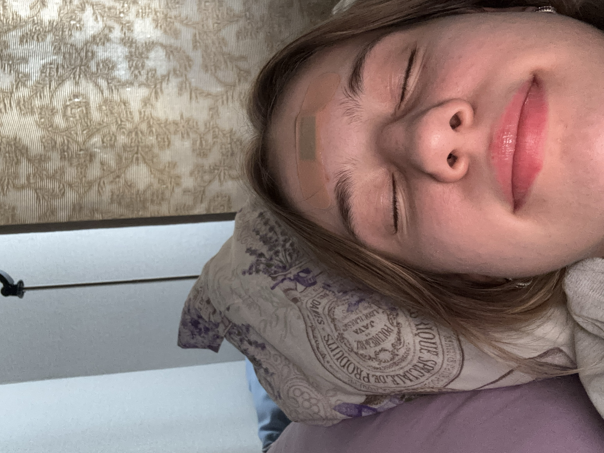 A young woman with closed eyes and a bandage on her forehead is lying down, possibly in a bed. She has blonde hair and is wearing a light-colored shirt. Her face is illuminated by soft lighting. The background includes a pillow with a floral pattern, a plain white wall, and a textured curtain or tapestry. There is no indication of the city of Lednevo, Russia, or any other specific location.